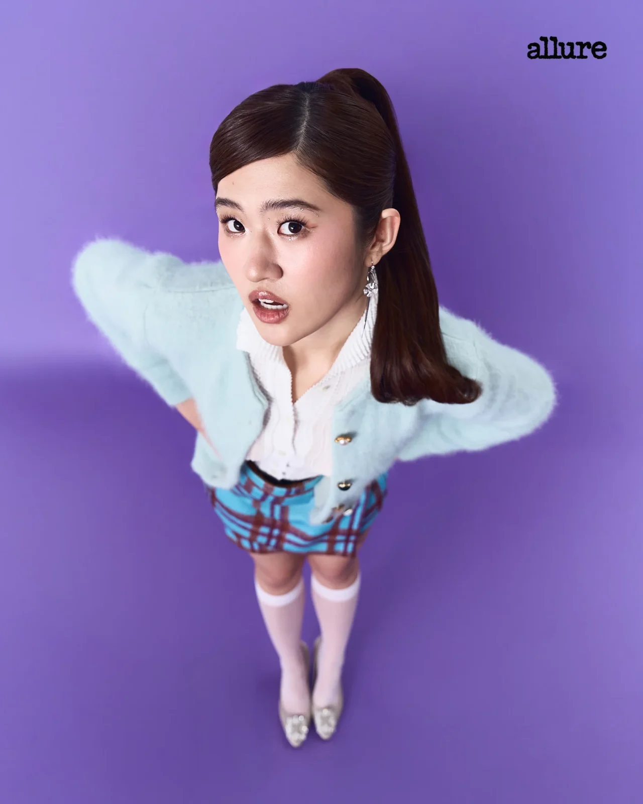 Young woman looking up at the camera against a purple backdrop, wearing a light blue cardigan, white blouse, plaid skirt and knee-high socks; Allure logo top-right.