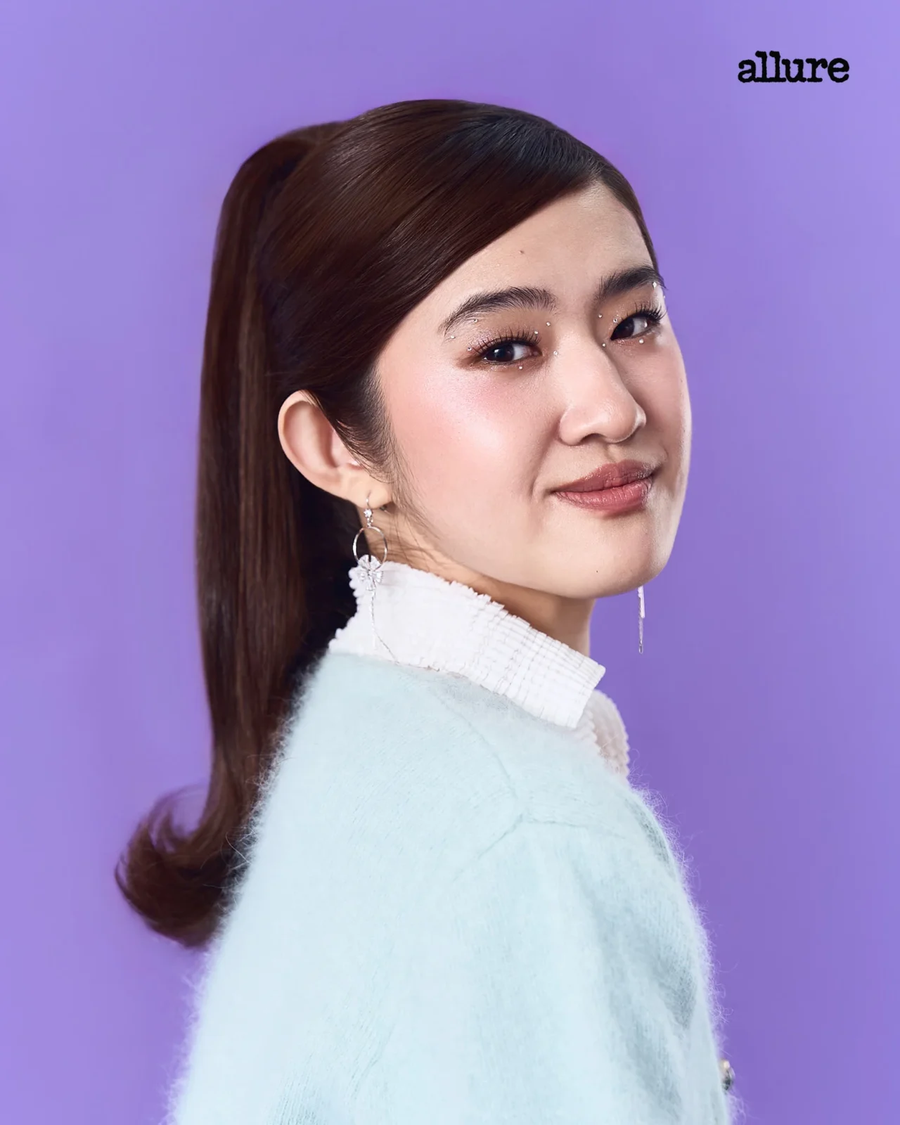 Side-profile portrait of a woman with long brown hair in a light sweater against a solid purple background; Allure logo in the top-right corner.