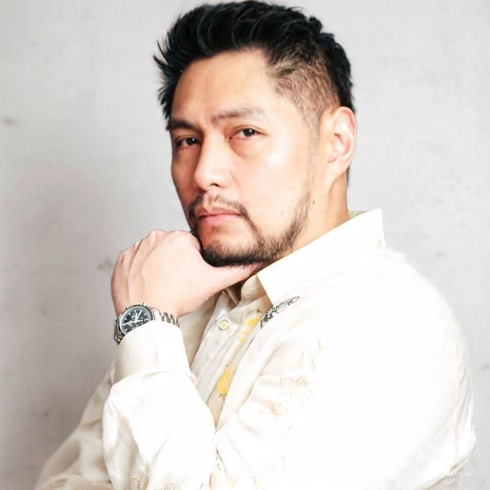 Man with short dark hair and beard, resting his chin on his hand, wearing a light beige shirt and a silver watch.