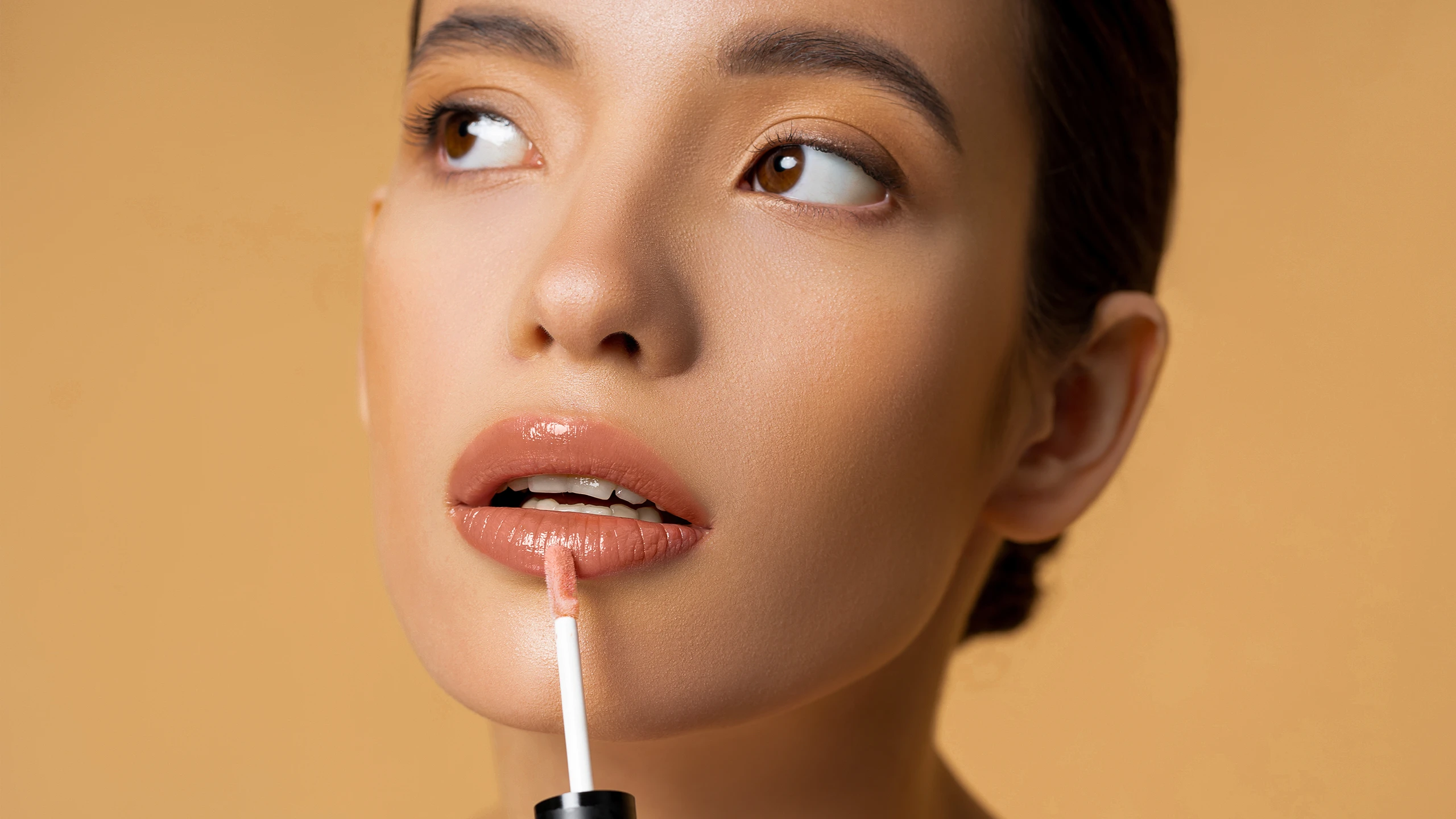 Close-up of a woman applying peach-toned lip gloss with a wand near her lips.