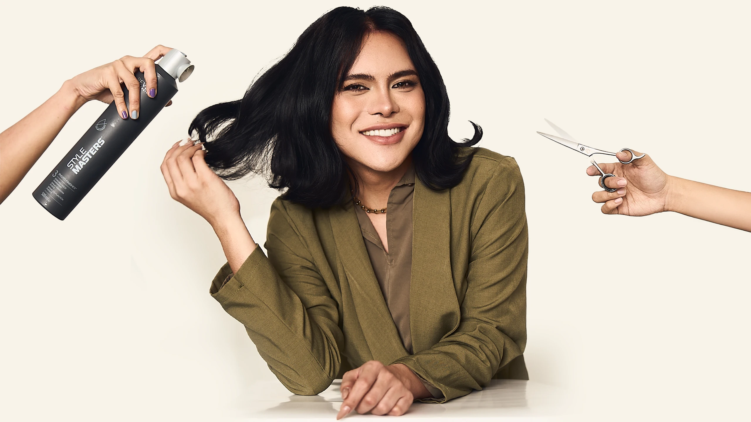 Smiling woman with dark hair while two hands hold a hairspray can and scissors near her hair, suggesting a styling session.