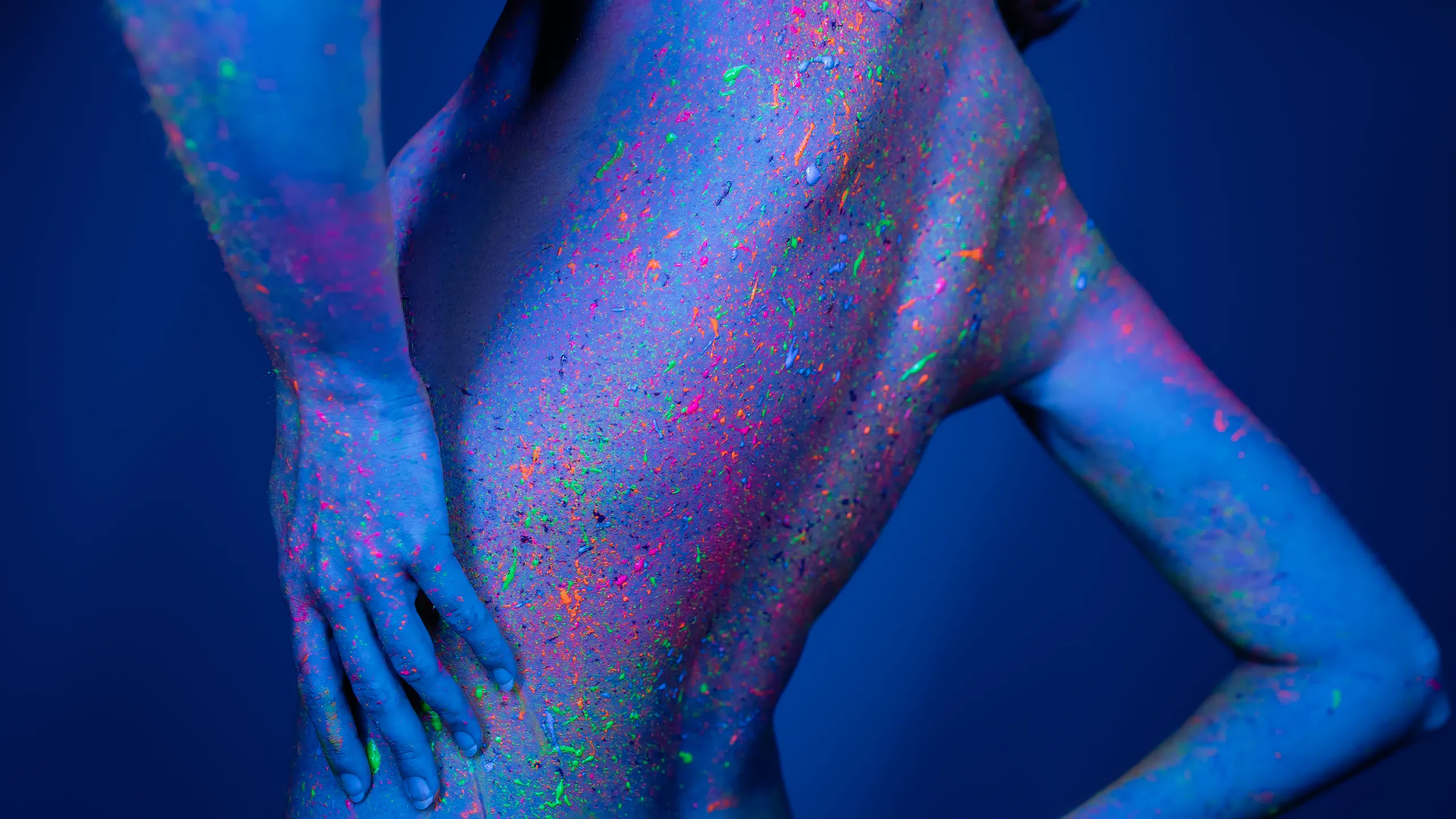 Back view of a nude person covered in neon paint under UV/blacklight, with splattered colors across the skin.