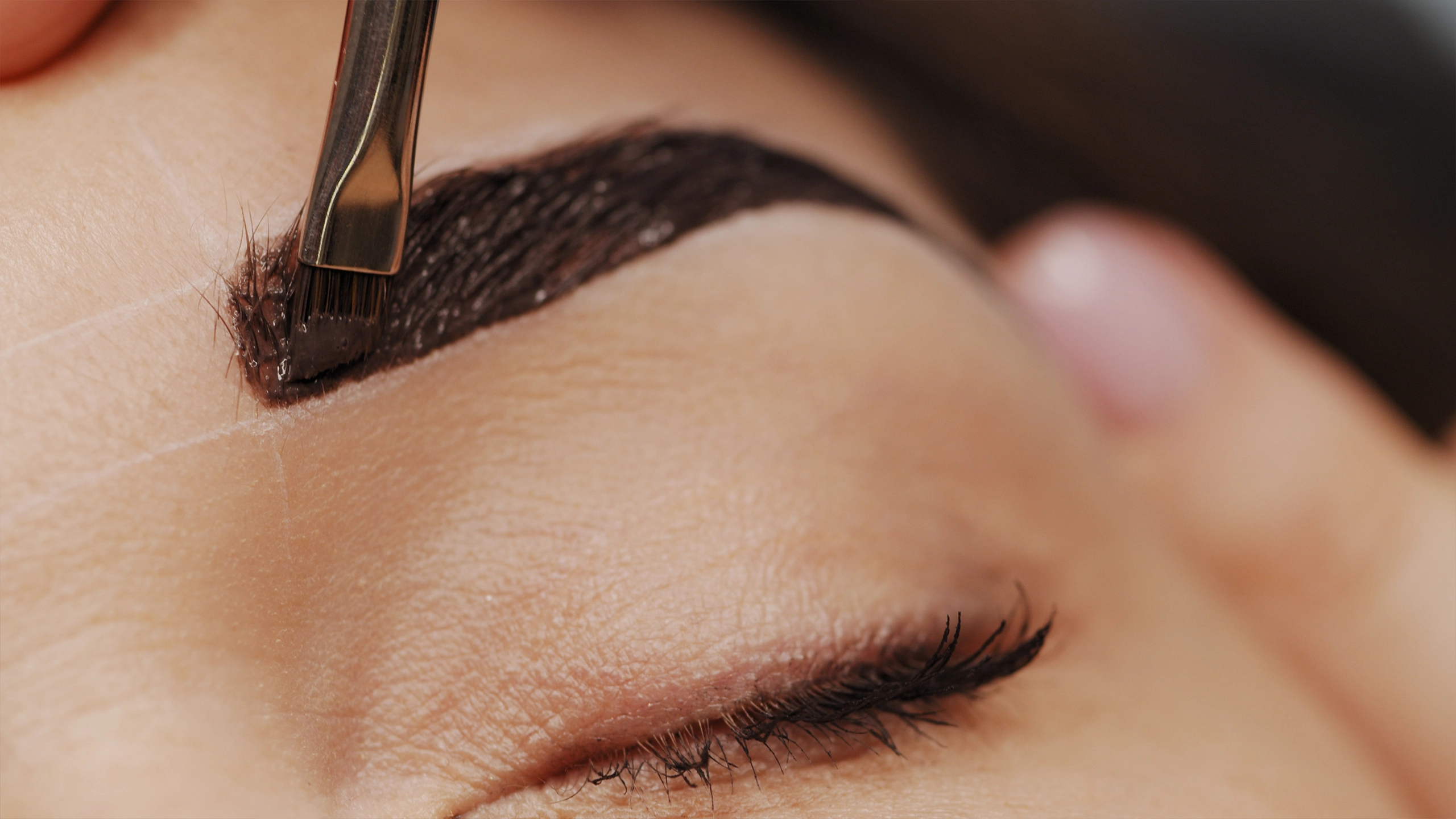 Close-up of a makeup artist applying dark brow pigment with a flat brush on a client's eyebrow during a cosmetic procedure.