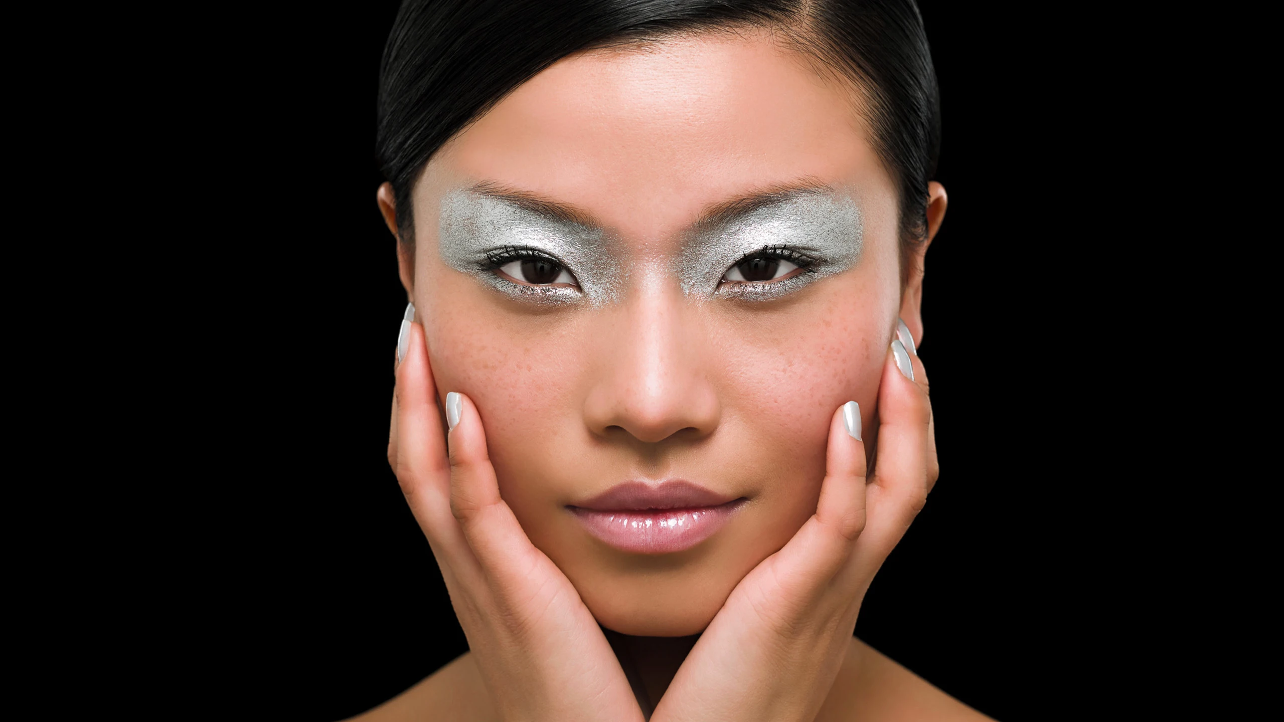 Close-up of a woman's face with silver glittery eyeshadow, gently cupping her cheeks with both hands against a black background.