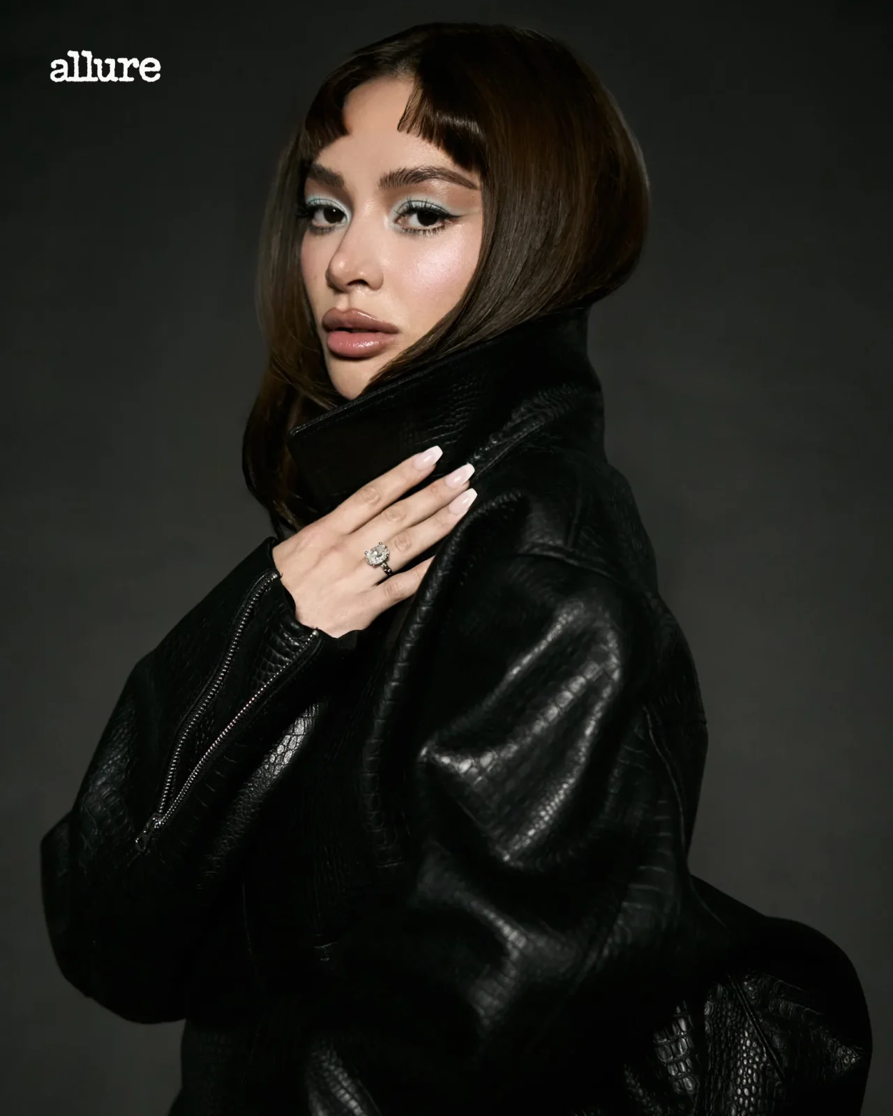 Portrait of a woman with bold makeup wearing a black leather jacket, posed against a dark studio backdrop, hand resting near her collarbone.
