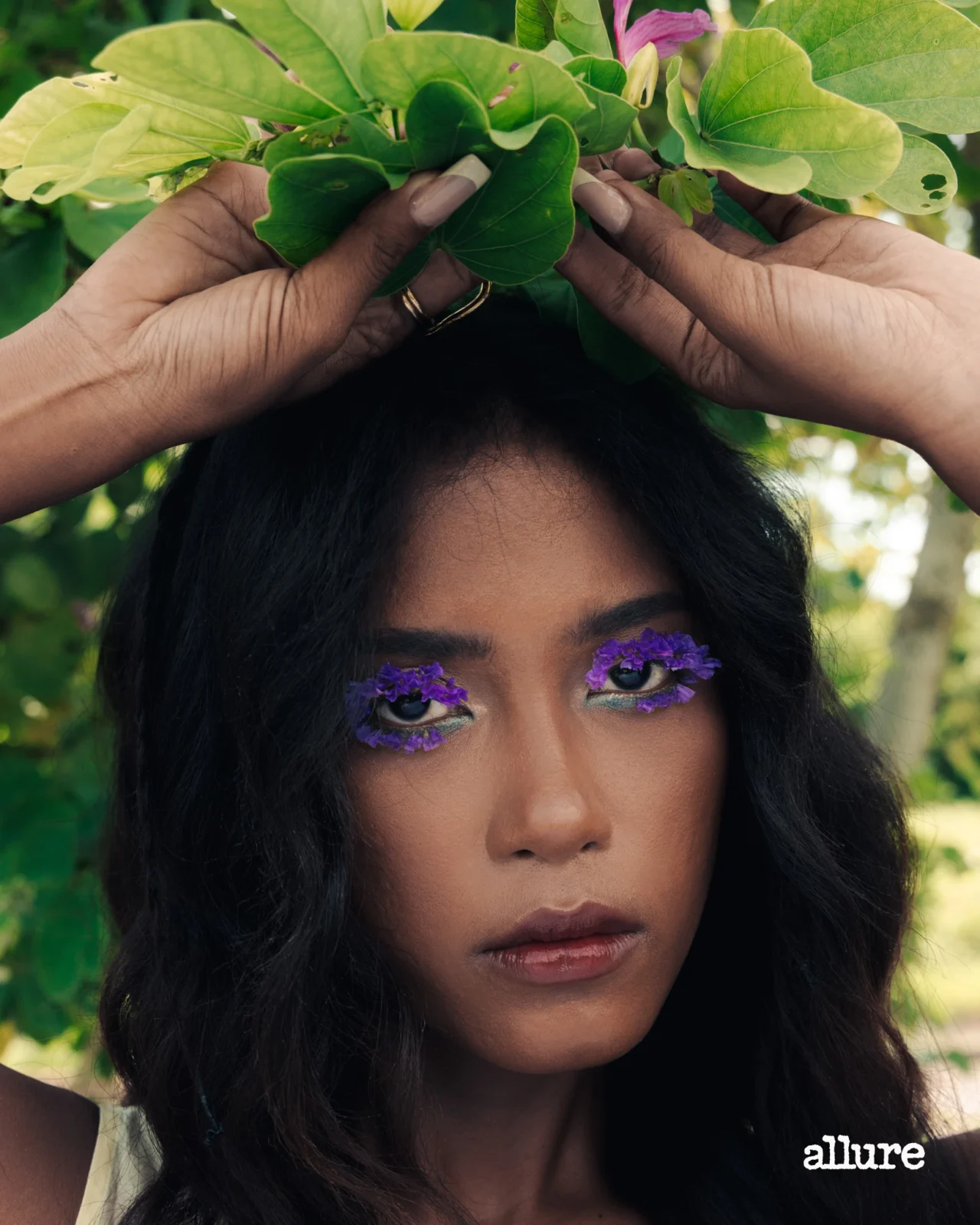 Close-up portrait of a woman with purple flower lashes, holding green leaves above her head outdoors.