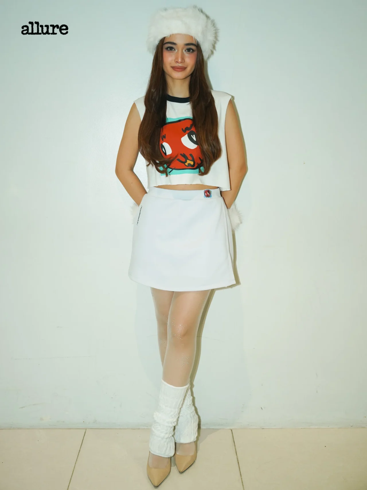 Young woman poses against a pale wall in a white cropped top with a colorful graphic, white skirt, fluffy white hat, and knit leg warmers; Allure logo in the corner.