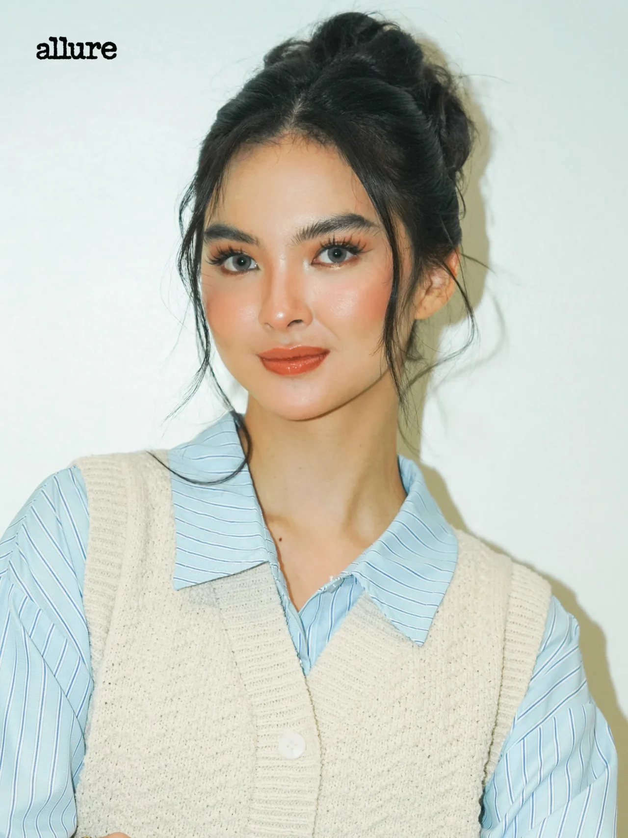Portrait of a woman with blue eyes and dark hair in a top bun, wearing a light blue striped shirt and beige knitted vest, looking at the camera.