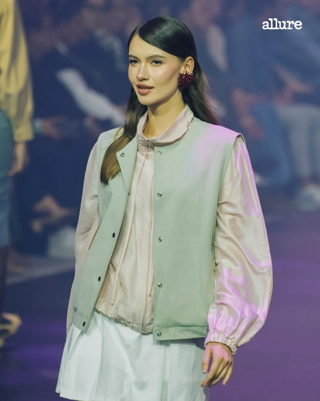 Fashion model wearing a pale pink blouse and mint vest, with a red flower earring, on a runway; Allure logo in the top-right corner.