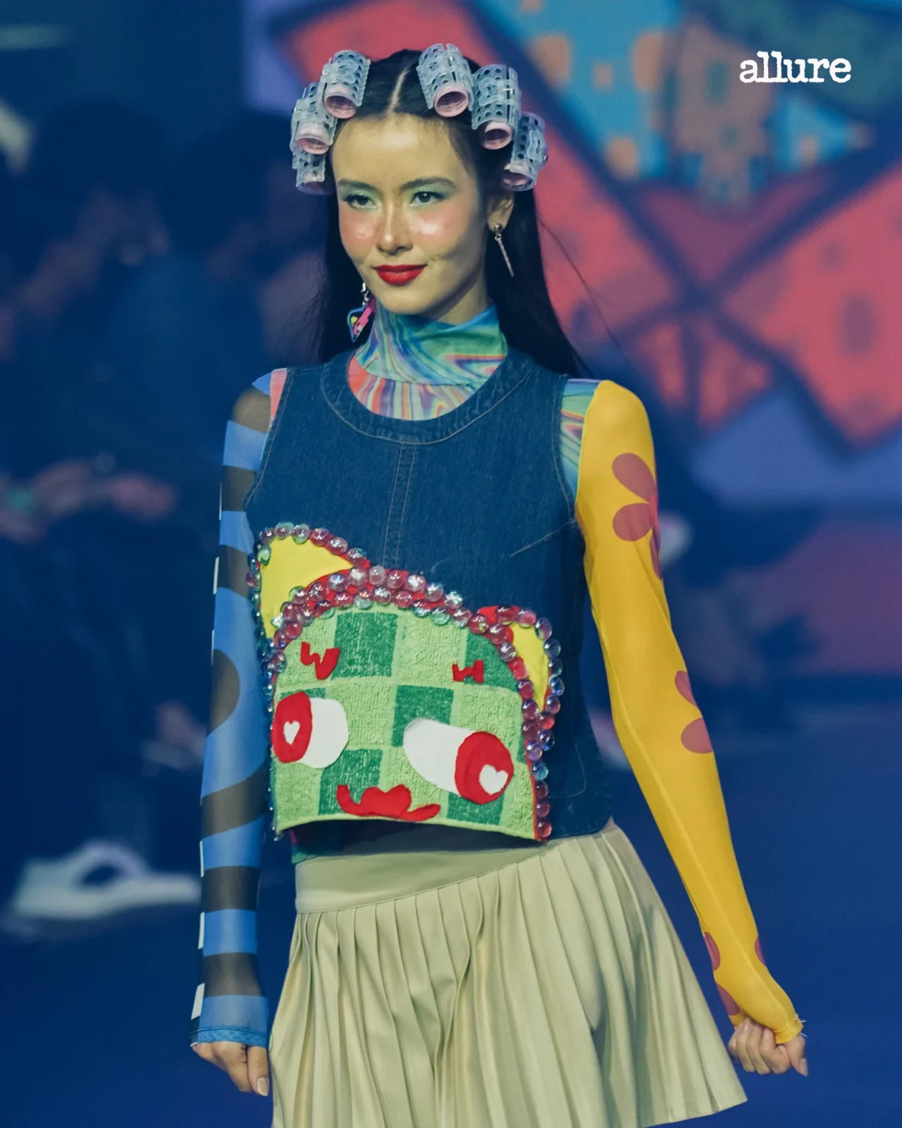 Model walks a fashion runway wearing a denim top with a bright, printed bib and colorful sleeves; hair in curlers, bold makeup, Allure logo in corner.