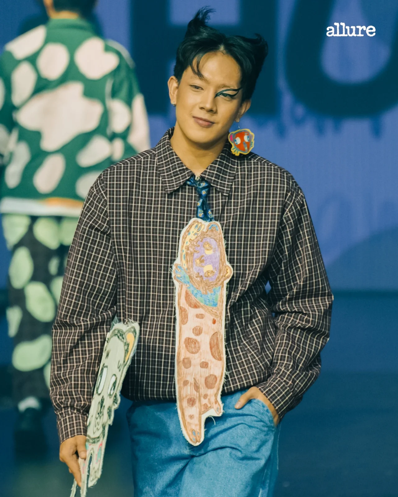 Person with short black hair and bold winged eyeliner wearing a black-and-white checkered shirt, holding large hand-drawn paper cutouts in front of a blue background with the Allure logo visible.