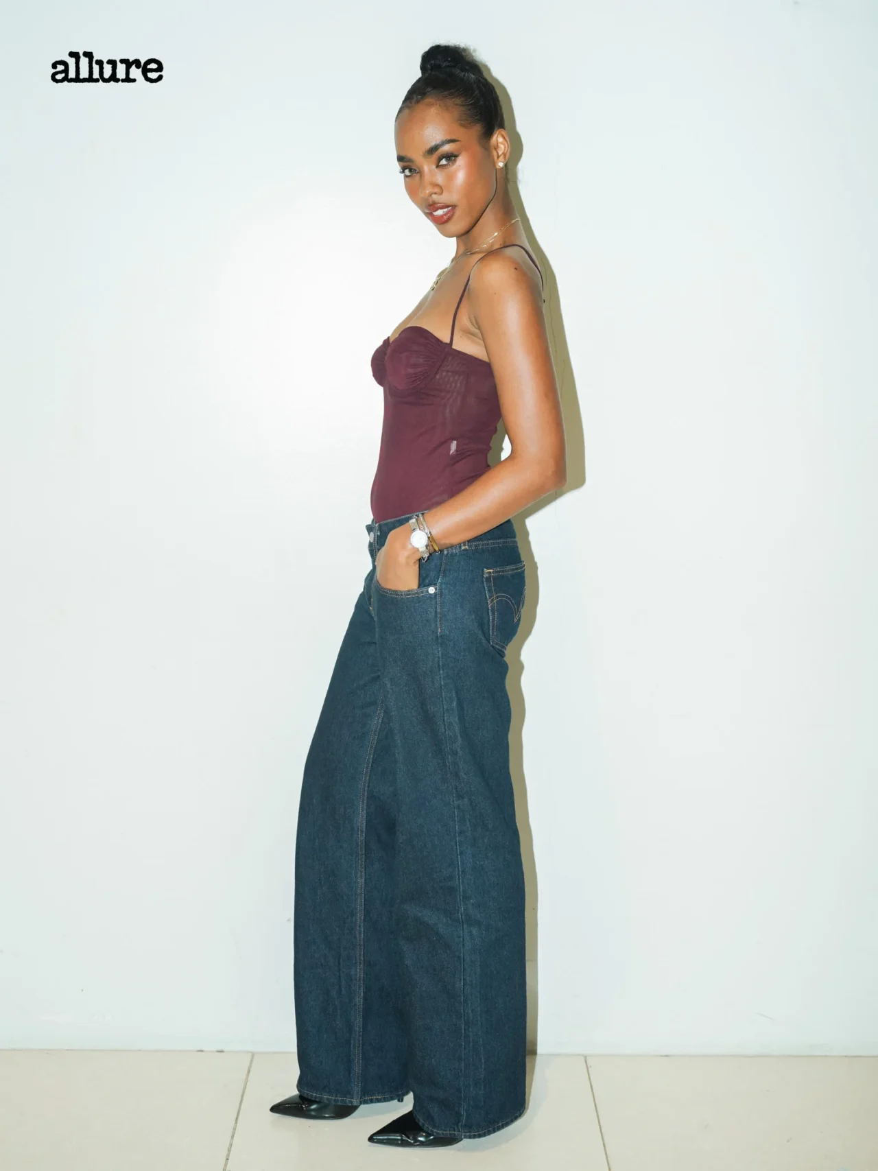 Model in a burgundy spaghetti-strap top and wide-leg jeans, posing in profile against a pale wall with the Allure logo in the top-left corner.