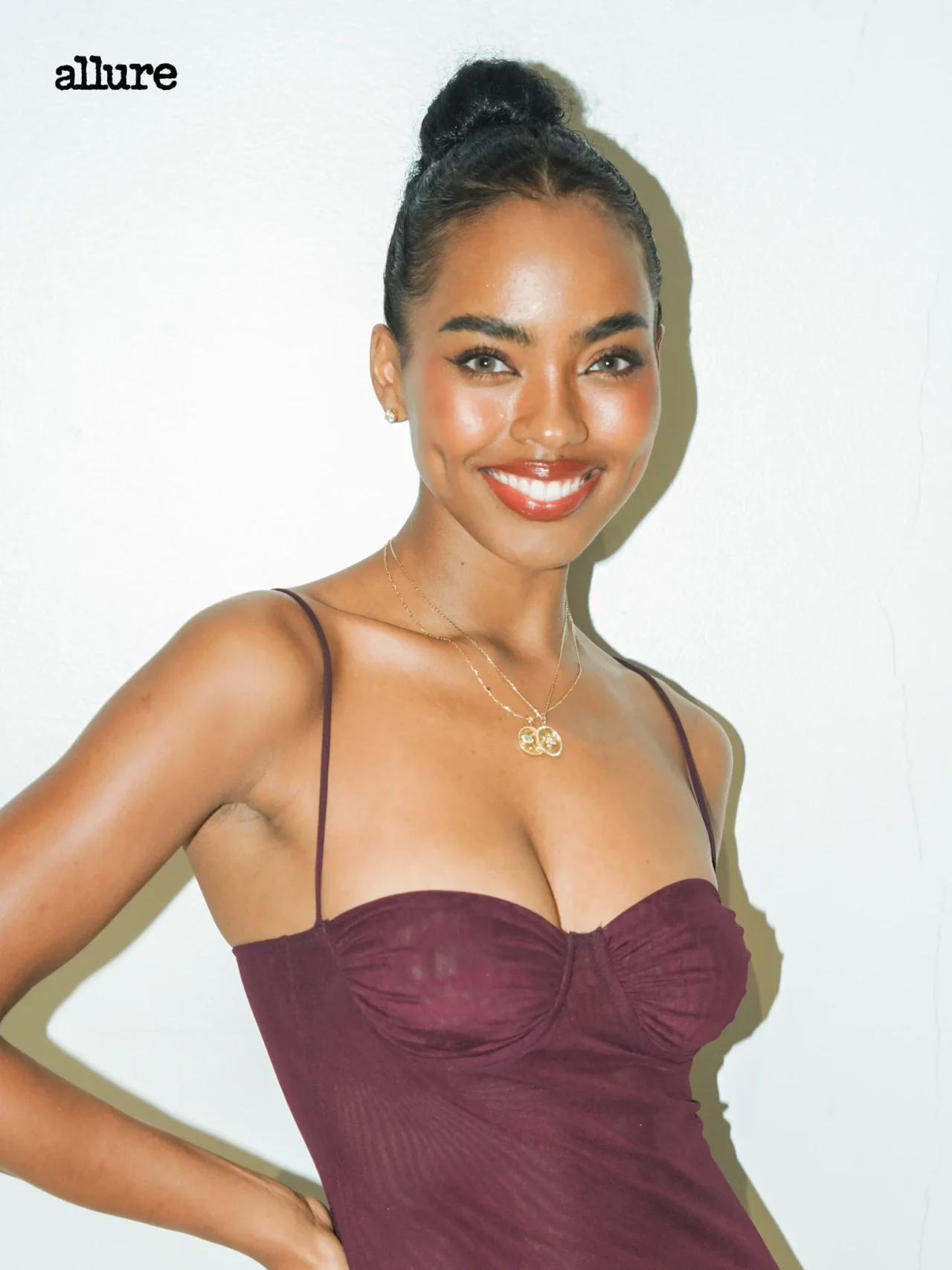 Smiling woman in a burgundy strapless dress with a gold pendant necklace, standing against a pale wall; Allure logo in the top-left corner.