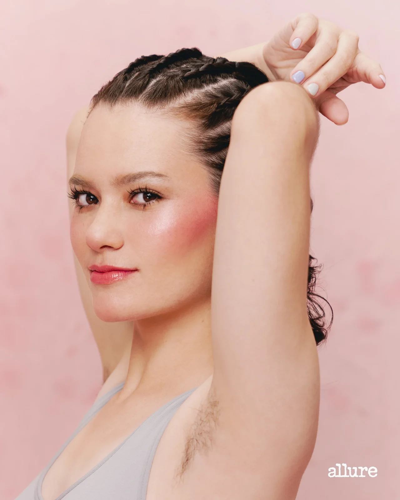 Portrait of a woman with braided hair and rosy makeup, arms raised against a pink backdrop (Allure logo bottom right).