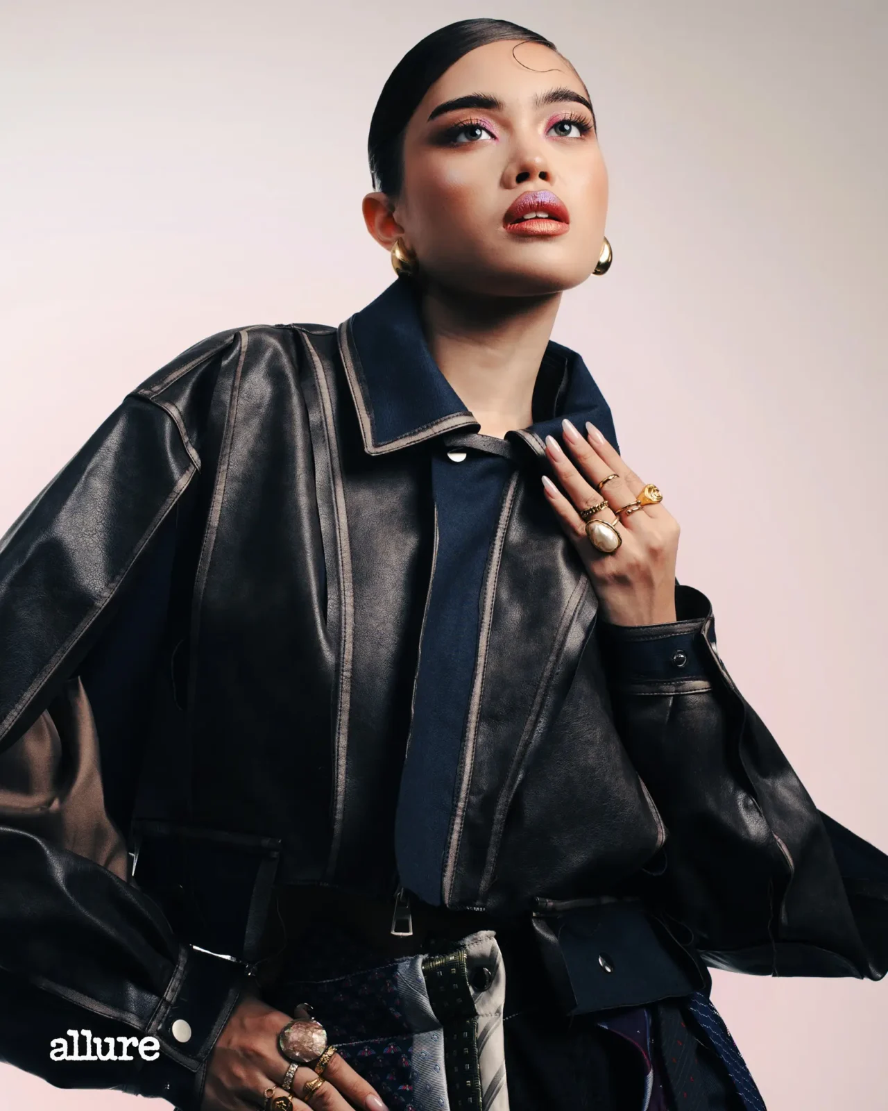 Editorial portrait of a woman in a black leather jacket with gold jewelry and bold makeup, looking upward against a pale backdrop.