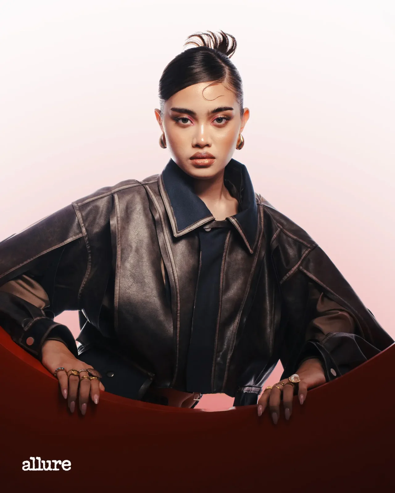 Fashion editorial portrait: model in a dark leather jacket, hands resting on a red surface, pale pink backdrop, Allure logo visible at bottom left.