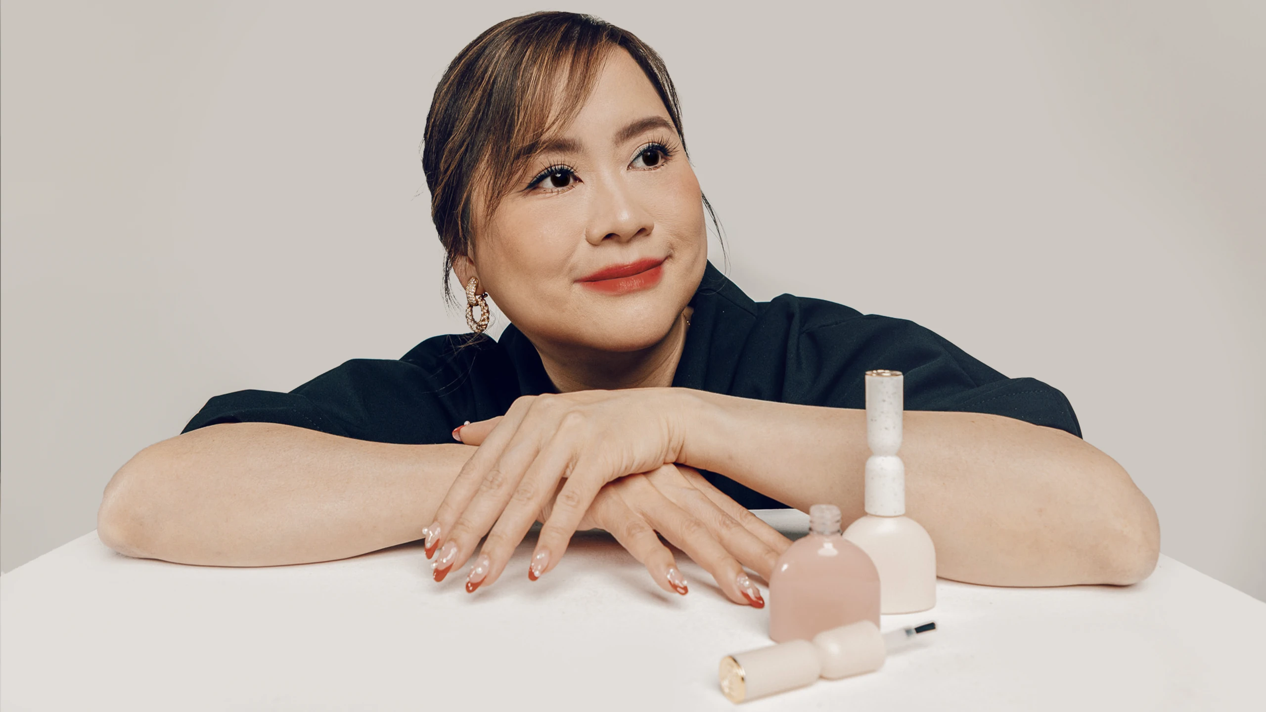 Woman resting her arms on a white table with nail polish bottles and makeup products nearby, looking to the left.