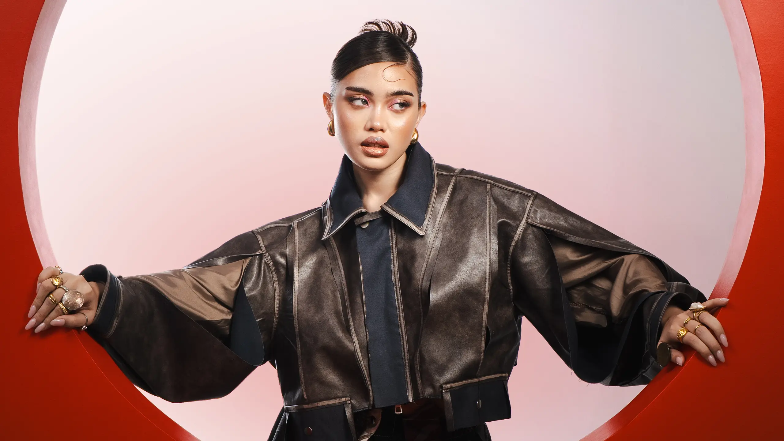 Model wearing a shiny dark metallic oversized jacket, arms through a red circular opening against a pink backdrop.