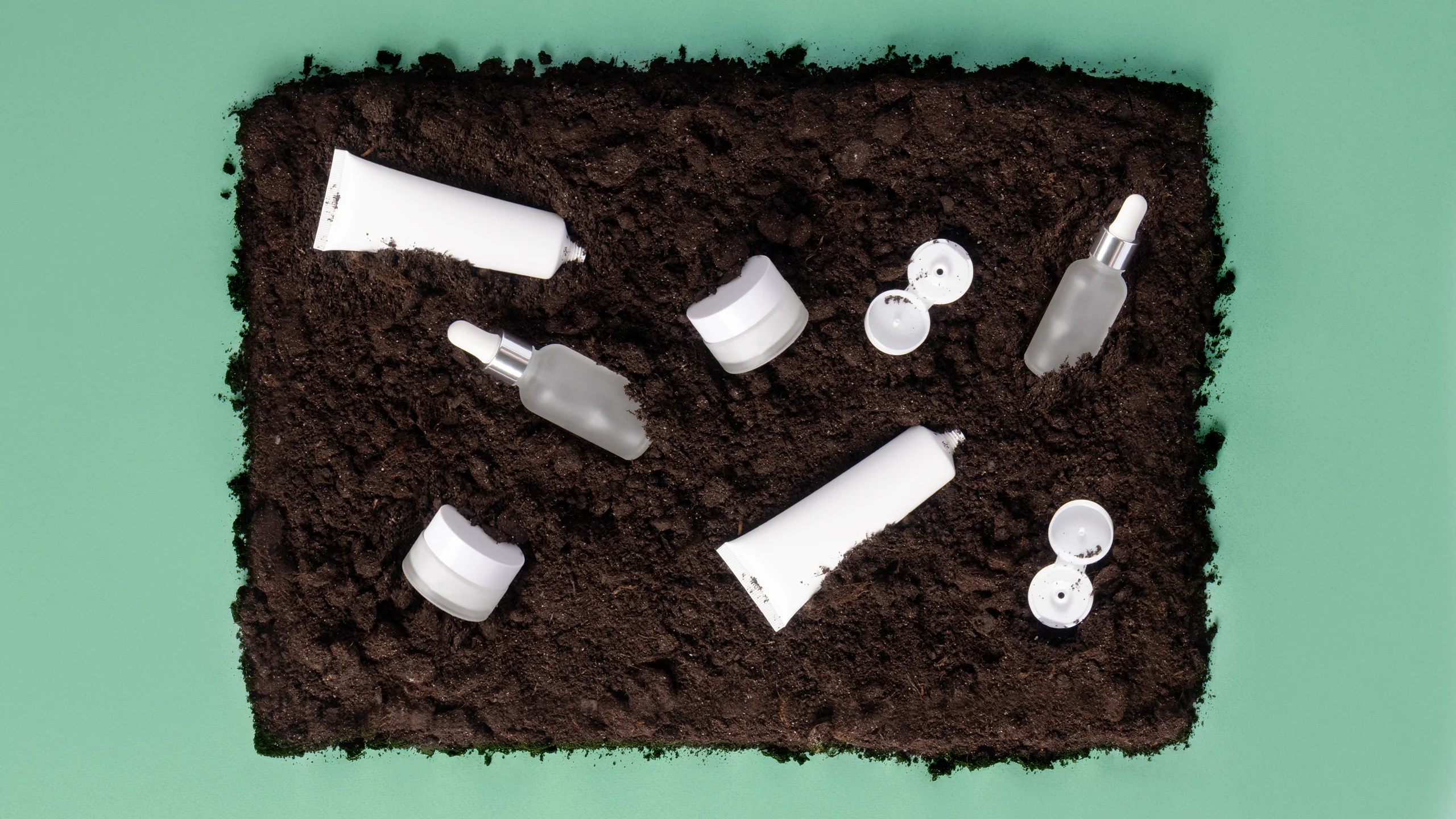 Assorted white skincare containers buried in dark soil on a green backdrop