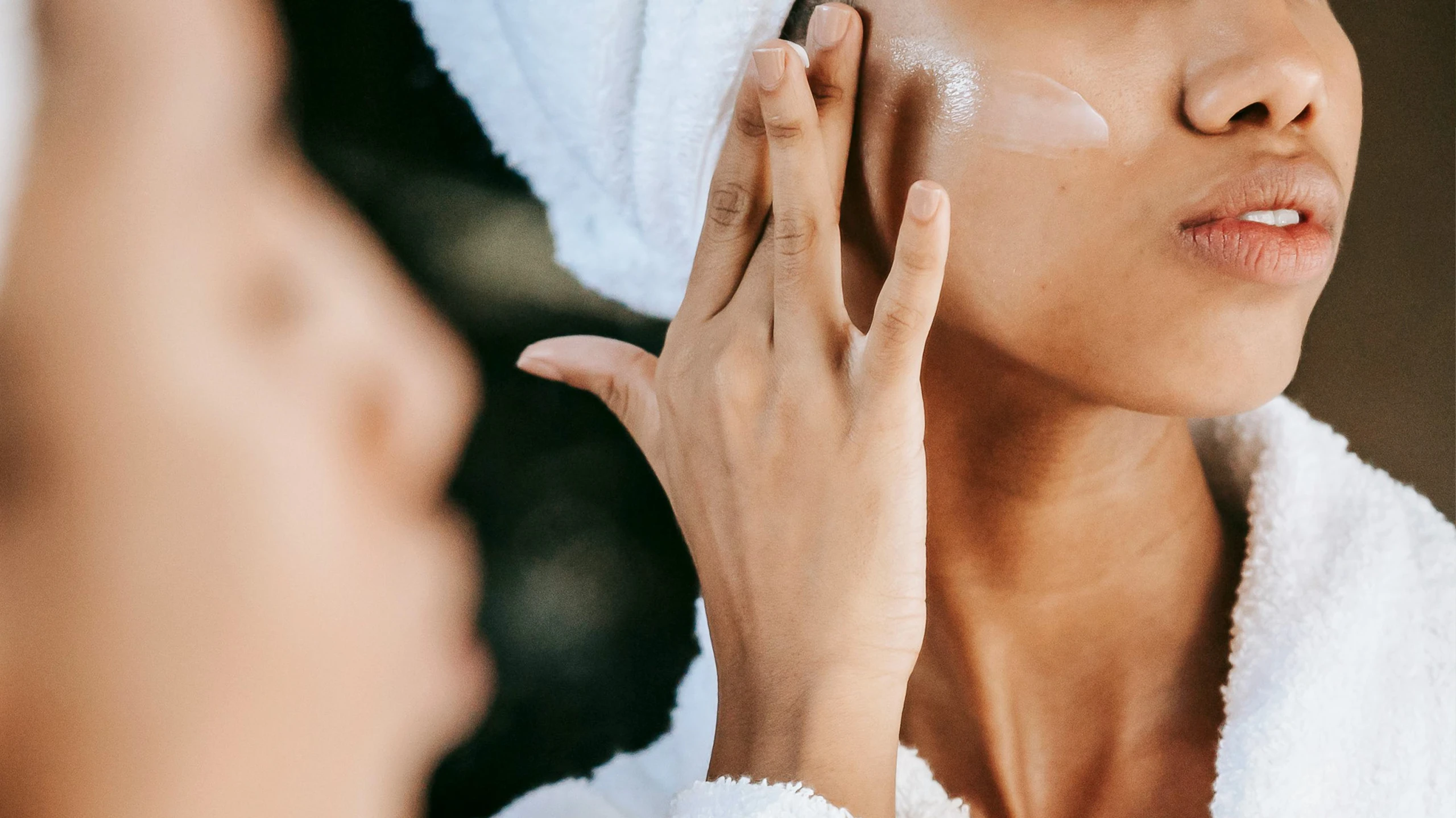 Close-up of a person applying cream under the eye with two fingers, wrapped in a white towel.