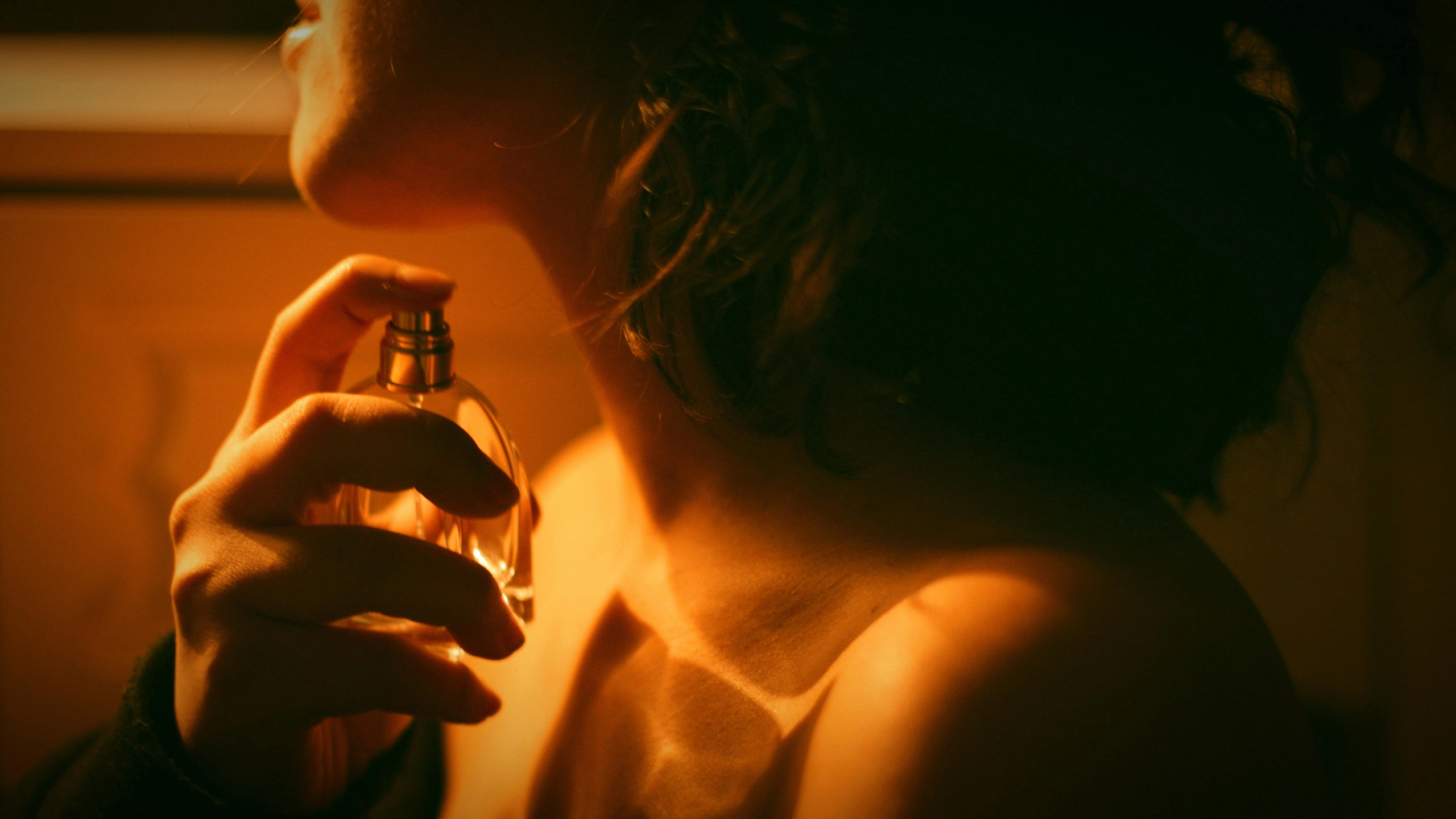 Close-up of a person applying perfume from a glass bottle, in warm orange lighting.
