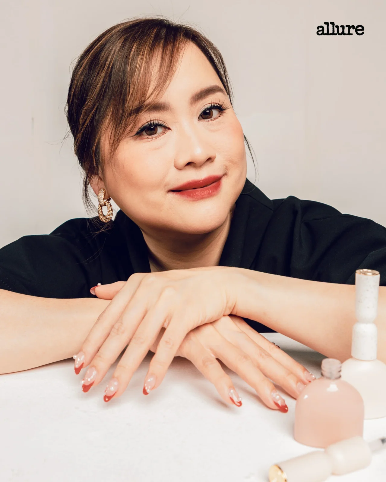 Close-up of a woman resting her hands on a table, displaying a glossy red French manicure with nail polish bottles nearby, Allure logo upper right.