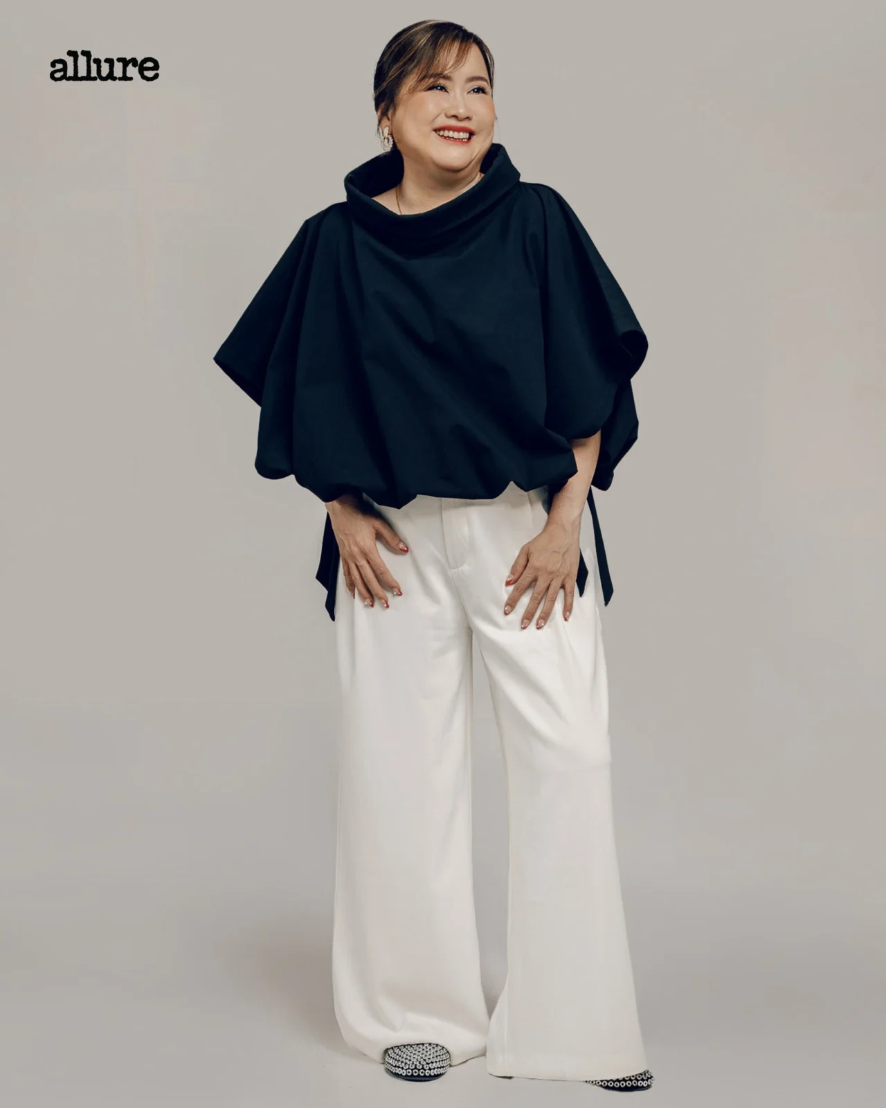 Smiling woman wearing a black draped poncho and white wide-leg pants, posing against a neutral gray backdrop with the Allure logo in the top-left corner.