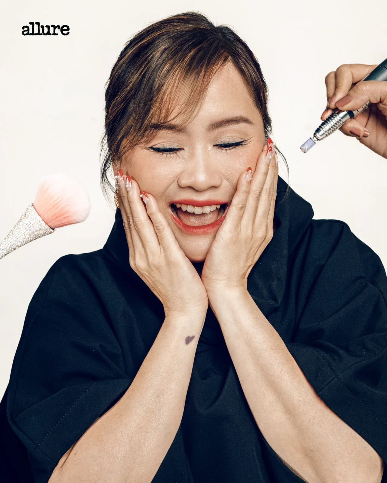 Joyful woman with eyes closed, hands on her cheeks as makeup is applied to her face by a tool held on the right.