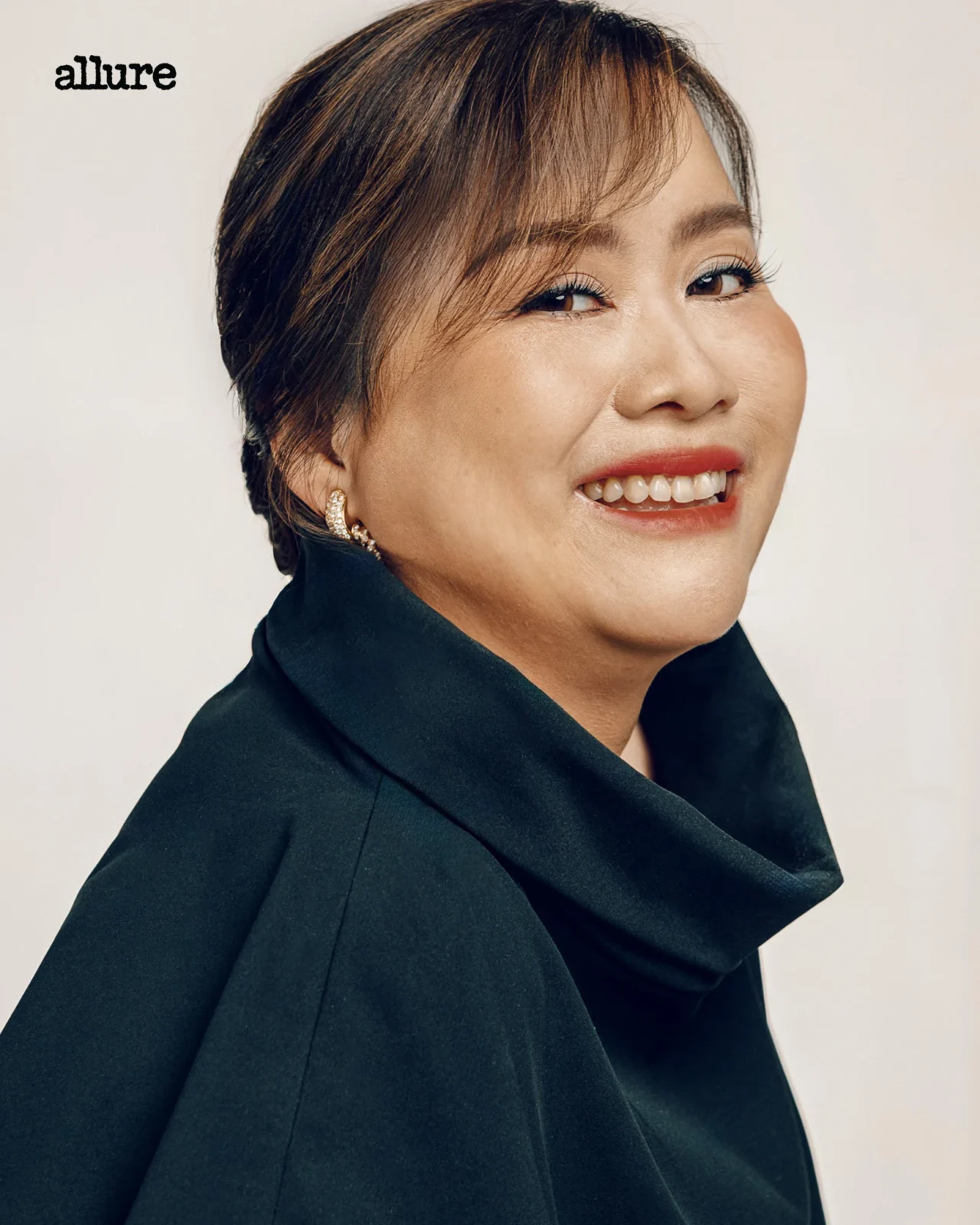 Close-up of a smiling woman with short dark hair, wearing a black blazer; 'allure' logo in the top-left corner.