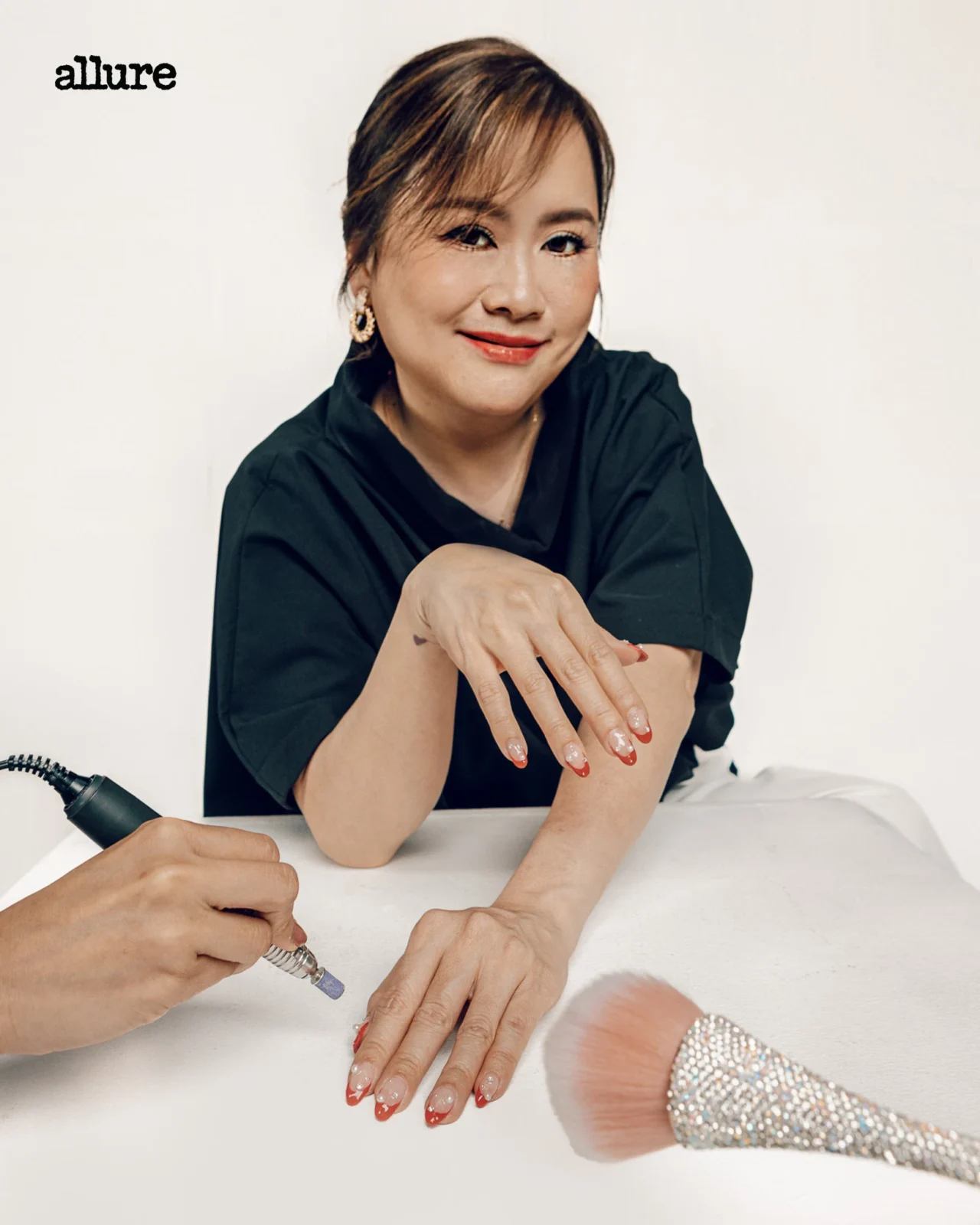 Smiling woman receives a manicure as a nail technician uses a drill on her nails; Allure logo in the top-left corner.