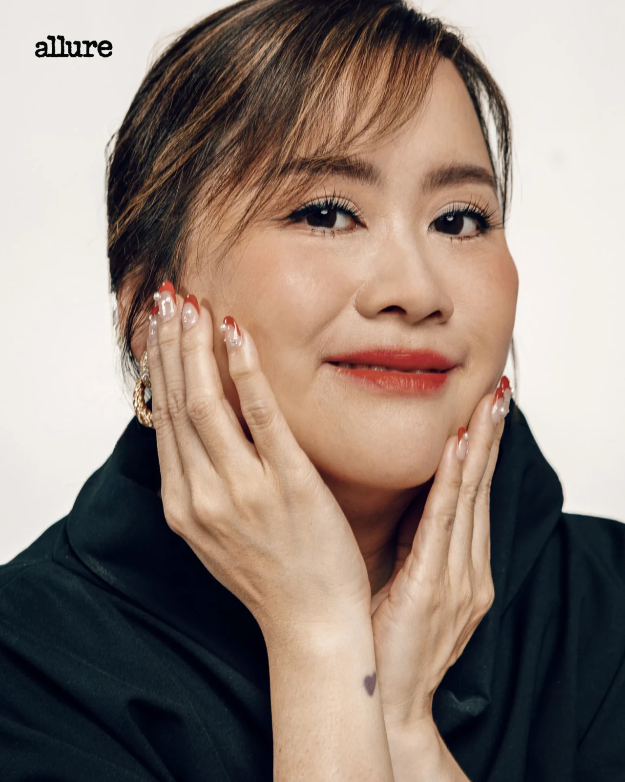 Close-up portrait of a woman with makeup resting her face in her hands, wearing a black top, small heart tattoo on her wrist, Allure logo in the corner.