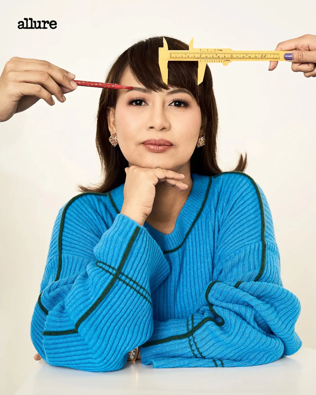 Woman in a bright blue knit sweater rests her chin on her hands while a red pencil touches her eyebrow and a yellow caliper spans her forehead for a beauty shoot (Allure logo visible).