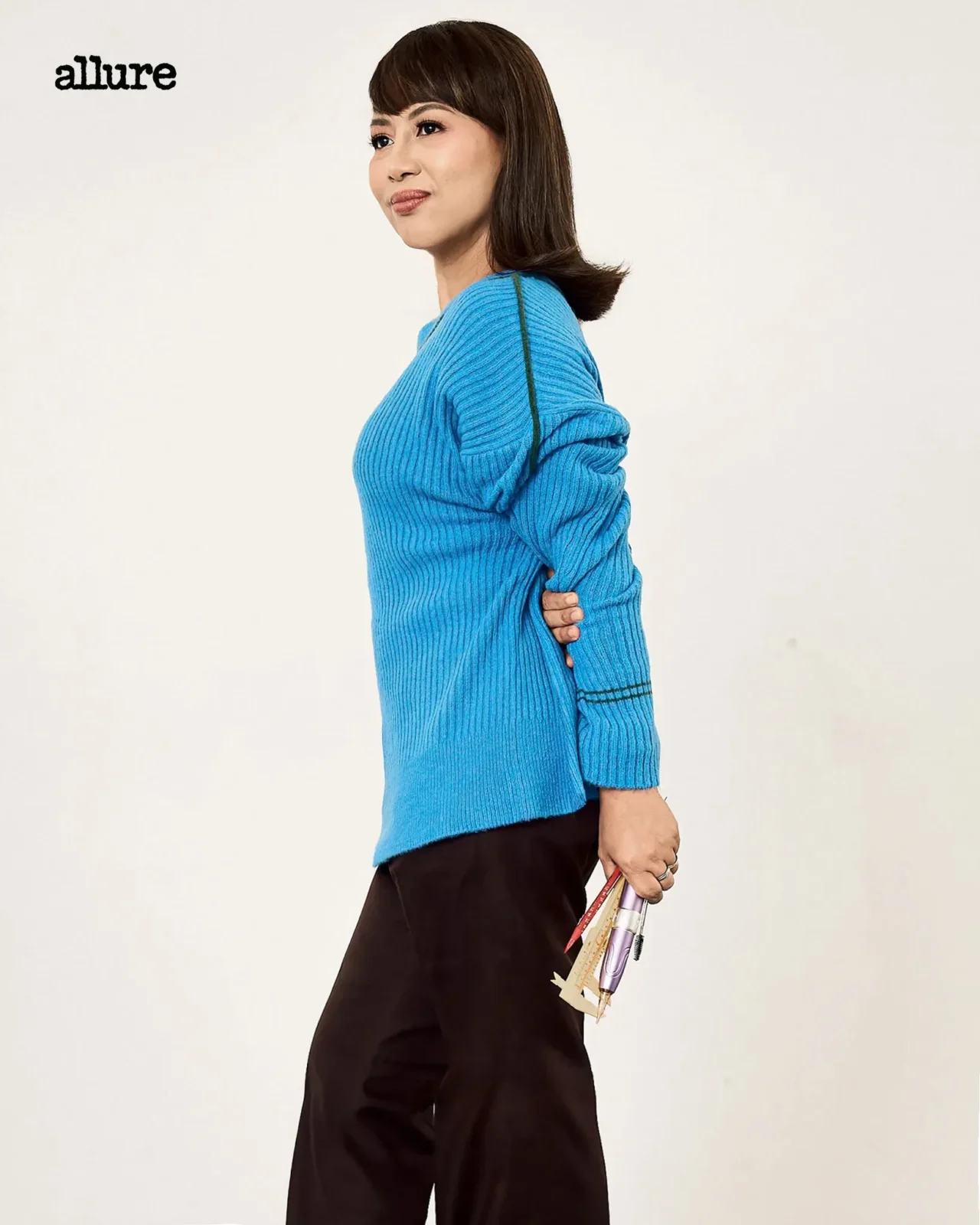 Side-profile image of a woman wearing a bright blue knit sweater, holding rolled papers in her right hand, against a pale background; the word 'allure' appears in the top left.