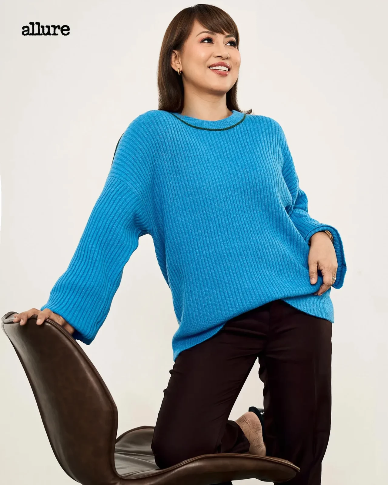Woman in a bright blue ribbed sweater leans on a brown chair, smiling against a light background with 'allure' logo in the corner.