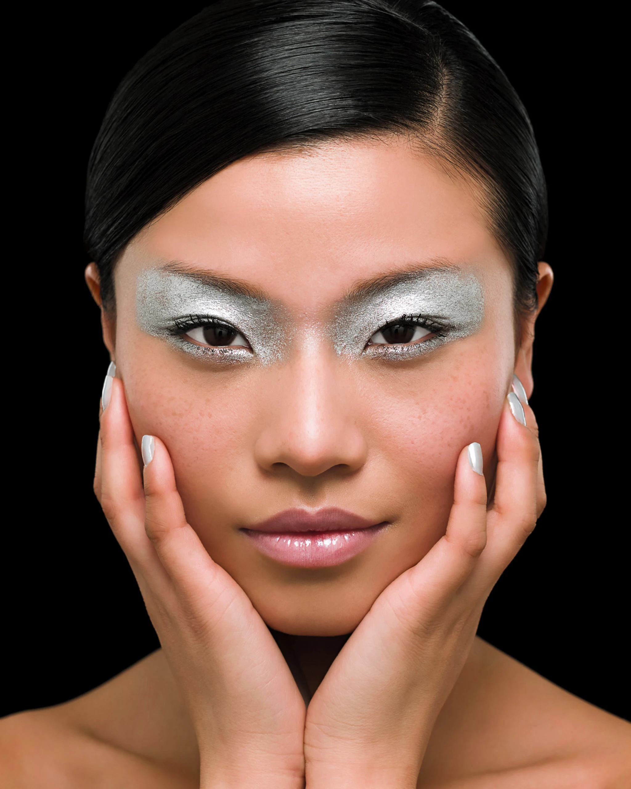 Close-up of a woman's face with metallic silver eyeshadow, hands on cheeks, black background.