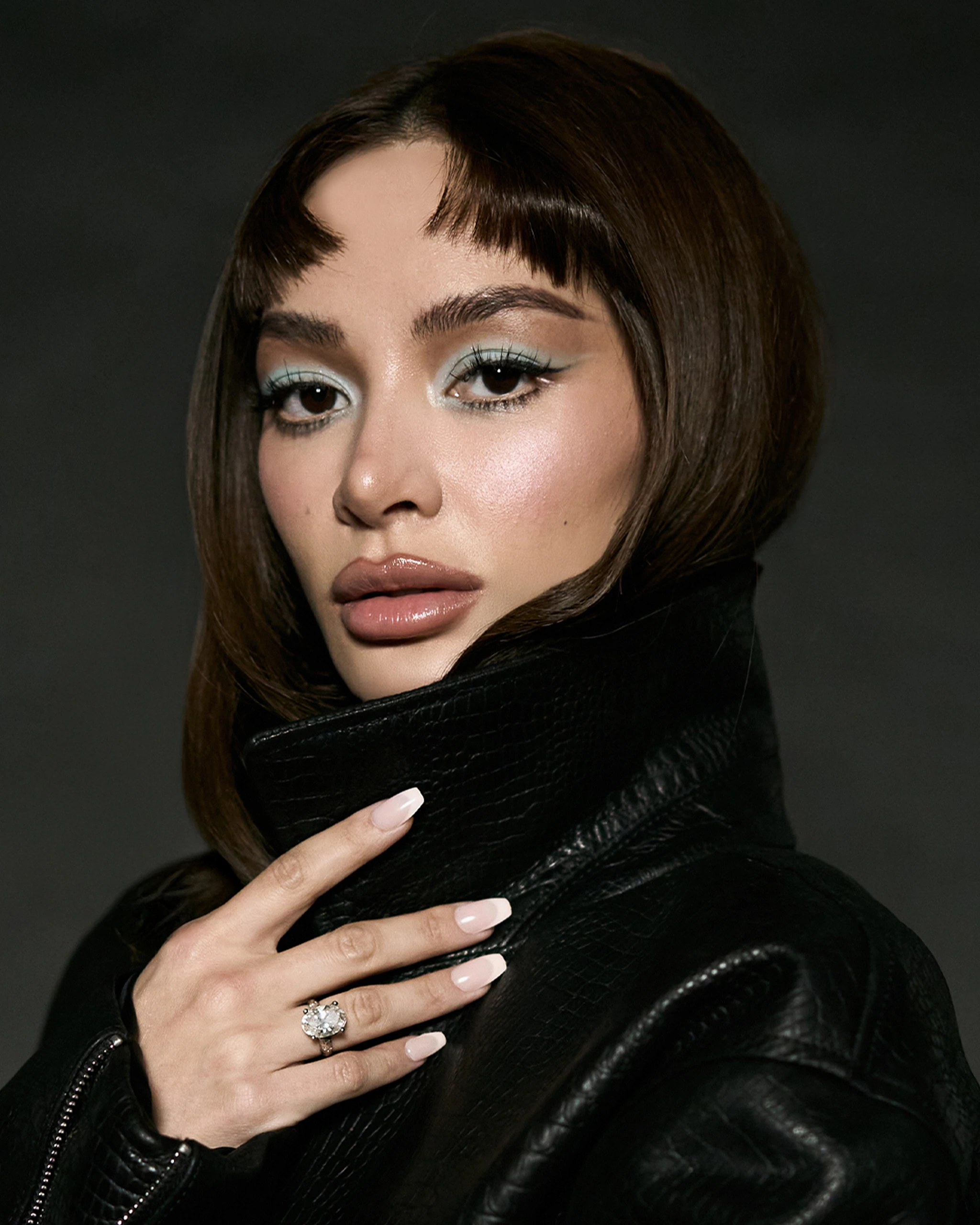 Close-up of a woman with bold makeup and glossy lips wearing a black leather jacket, hand resting near her chest.