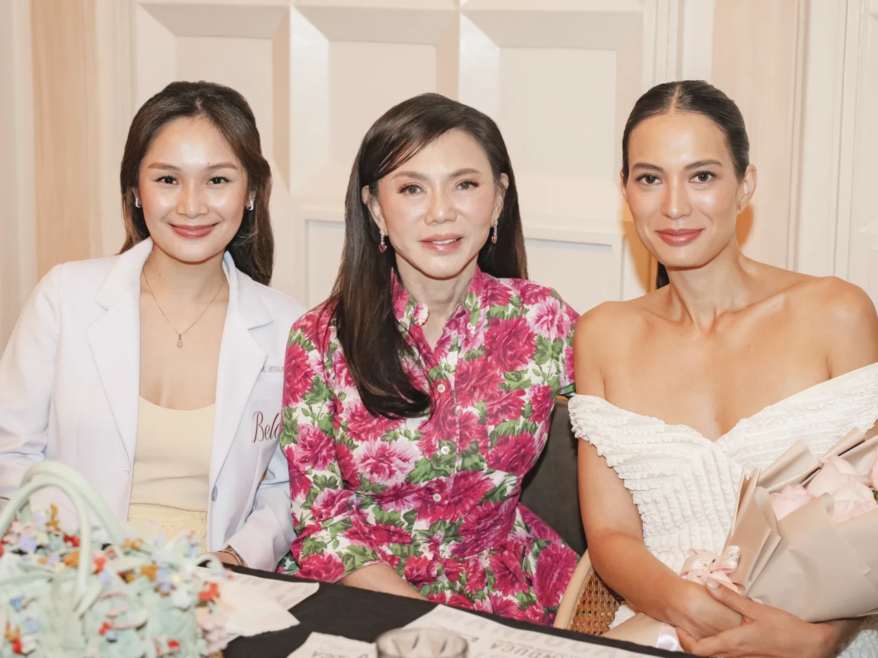 Three women pose together and smile for a photo at a social event; one in a white blazer, one in a pink floral dress, and one in an off-shoulder white dress holding a bouquet.