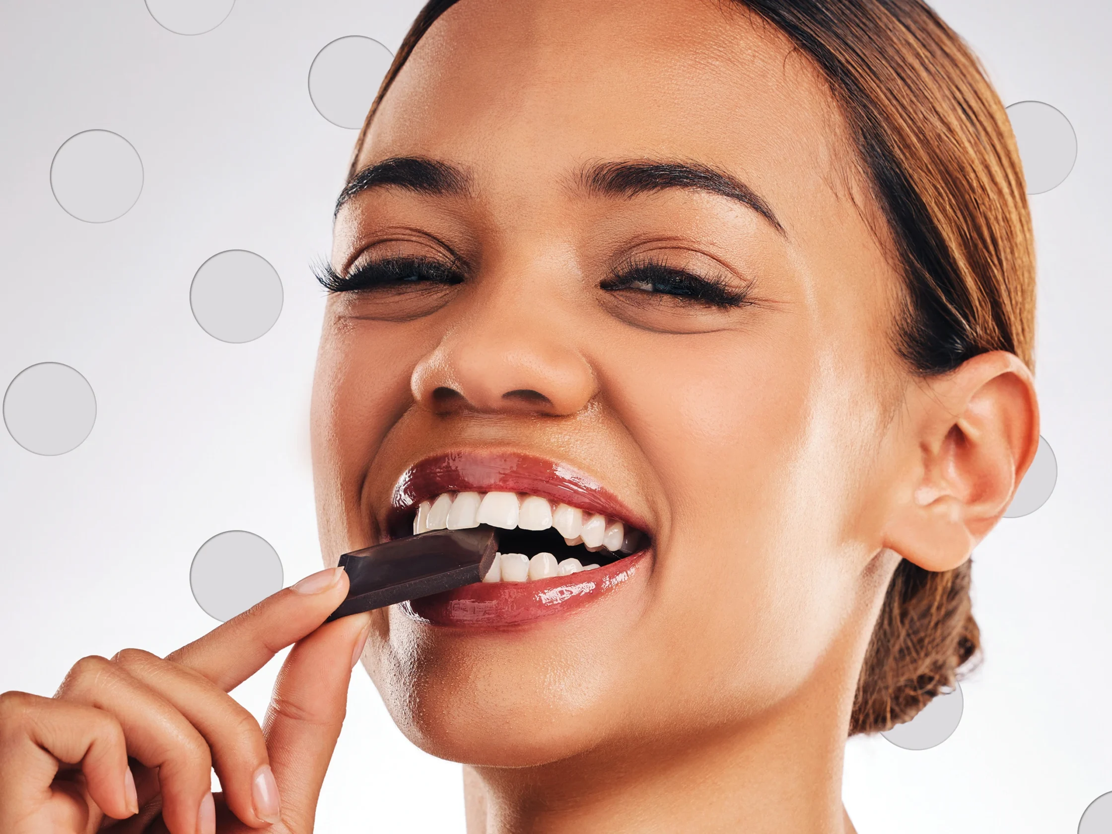 Close-up of a woman smiling as she applies red lipstick to her lips, showing glossy finish and white teeth.