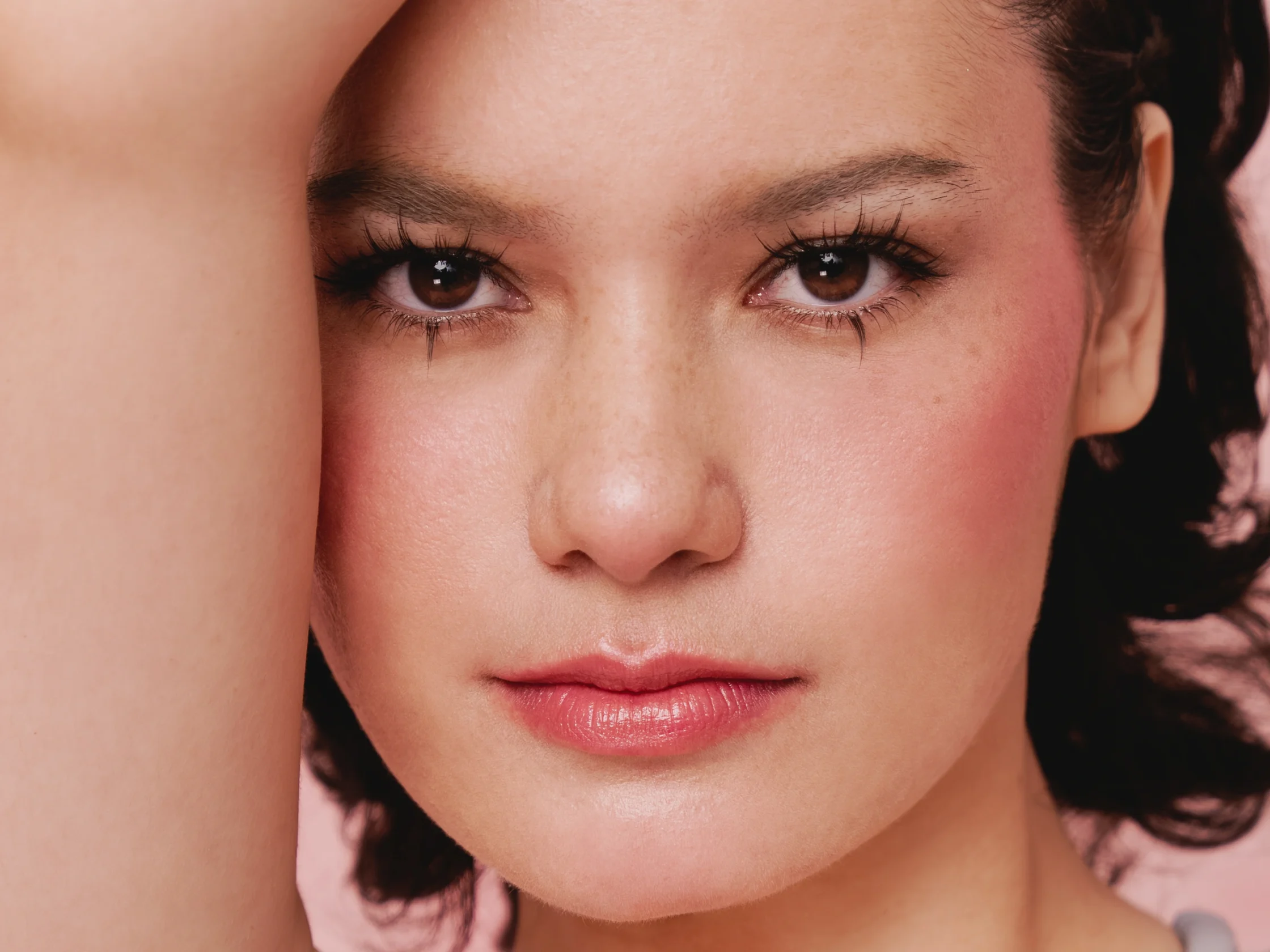 Close-up portrait of a woman with rosy cheeks and pink lips, one arm raised across her forehead as she looks at the camera.