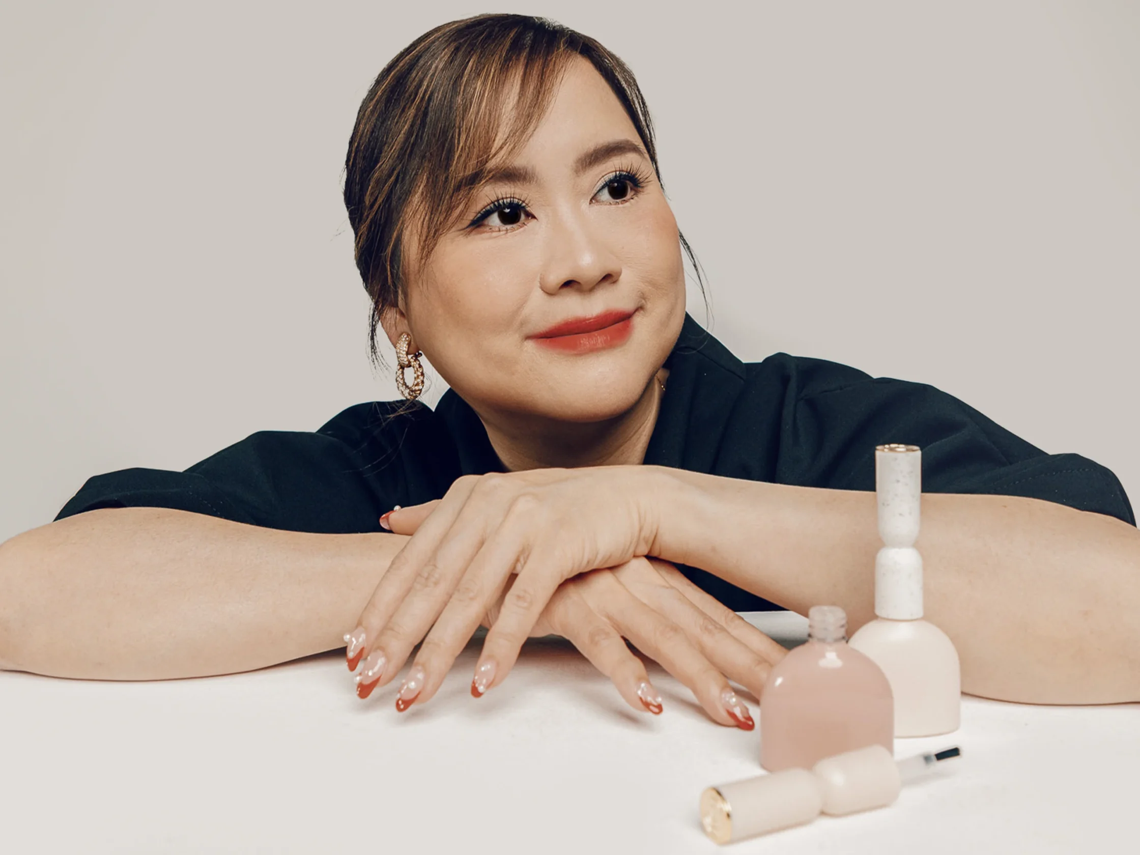 Woman resting her arms on a table during a manicure session, showing glossy red-tipped nails and several polish bottles nearby