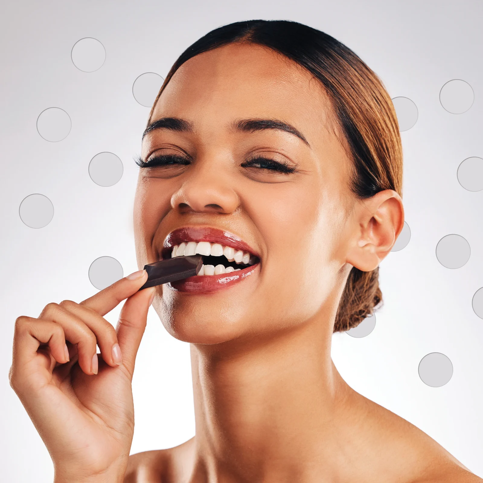 Close-up of a smiling woman biting into a piece of dark chocolate against a light background with circular patterns