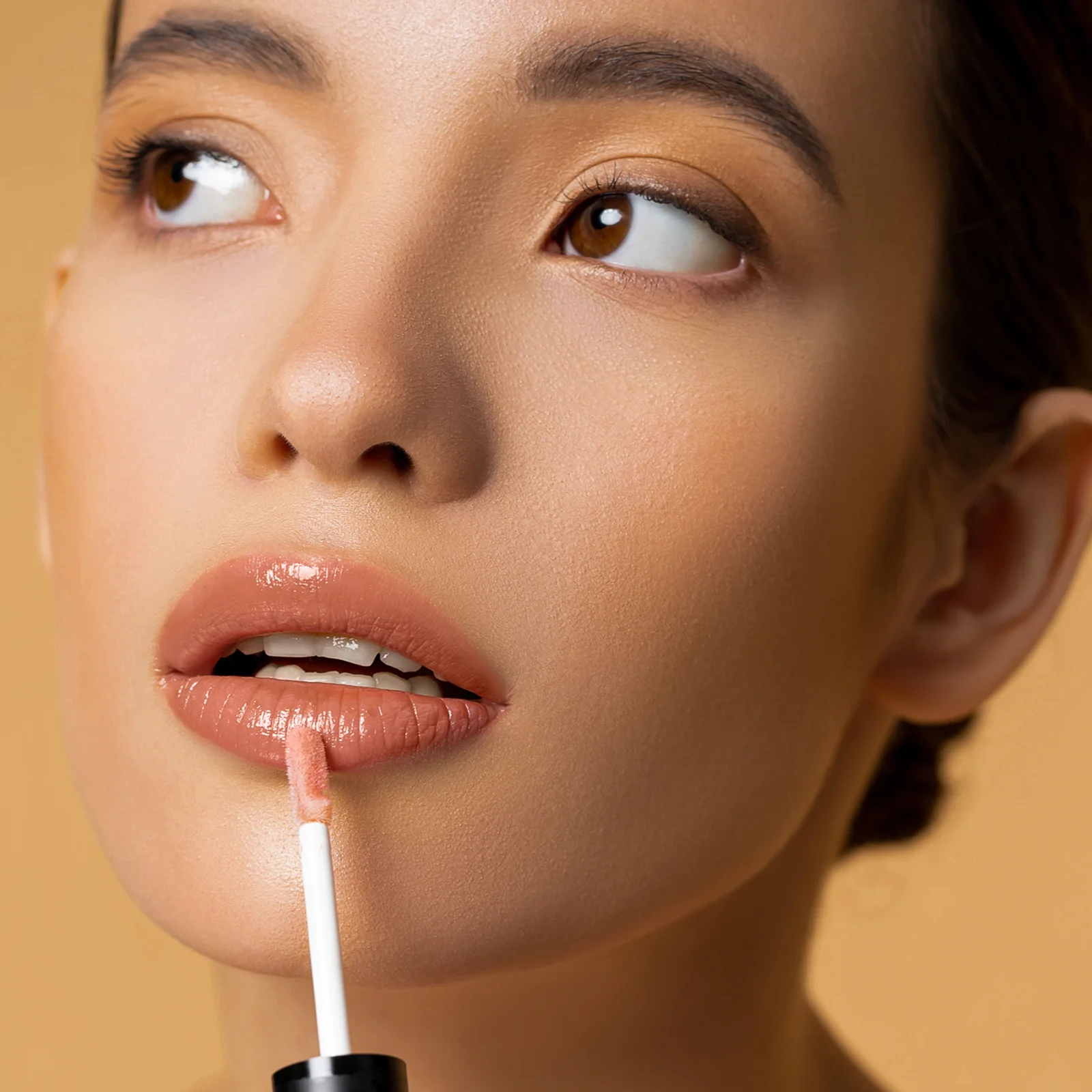 Close-up of a woman applying nude lip gloss with a wand, focusing on glossy lips and smooth skin