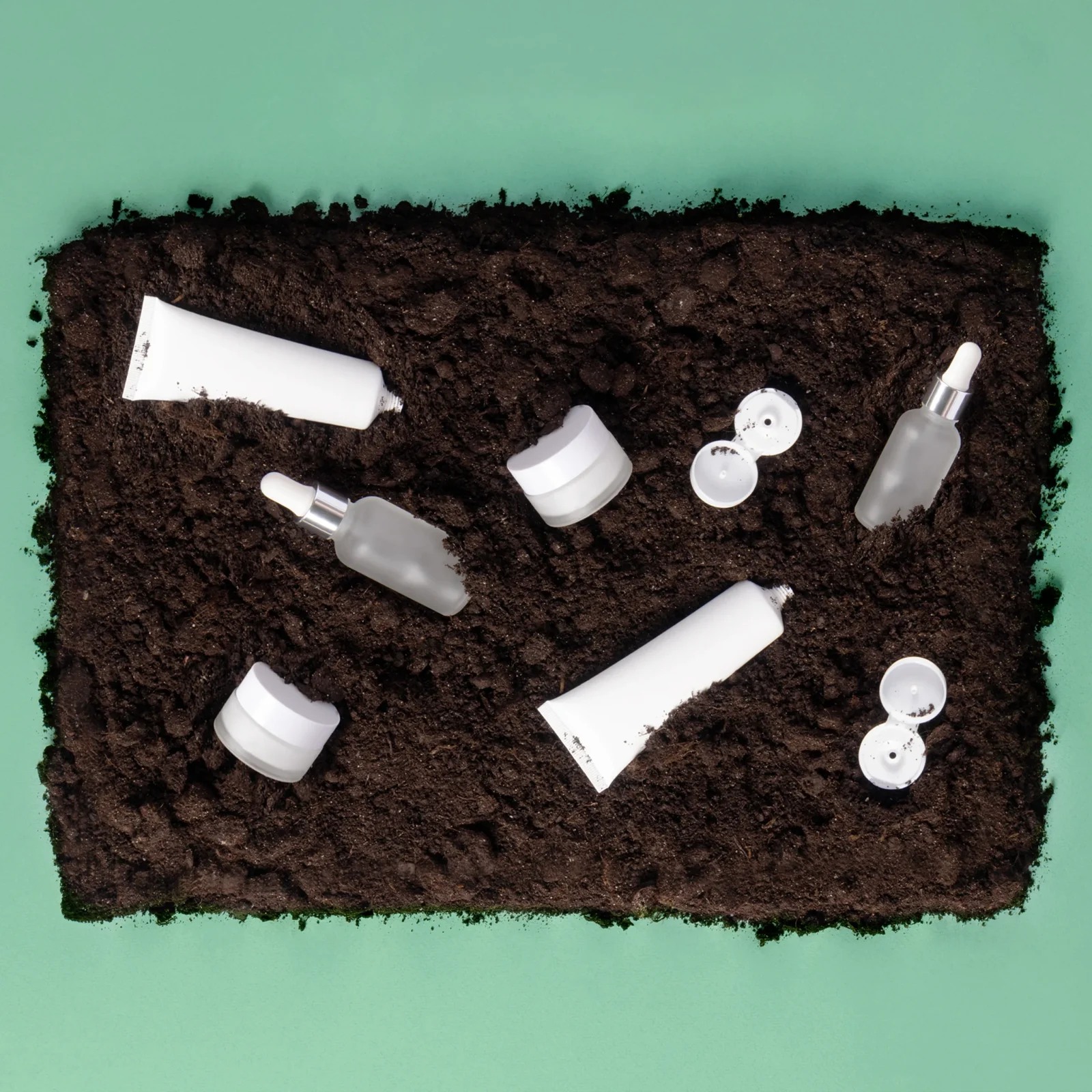White skincare containers (tubes, jars, droppers) scattered on dark soil inside a green tray.
