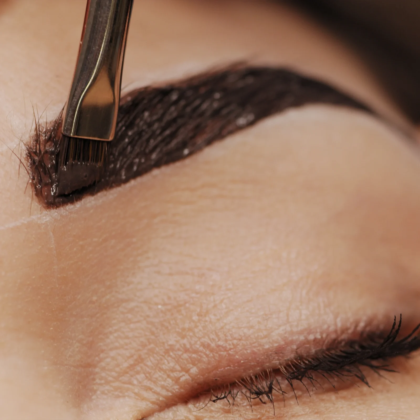 Close-up of a makeup brush applying dark brow product to an eyebrow for shaping and tinting
