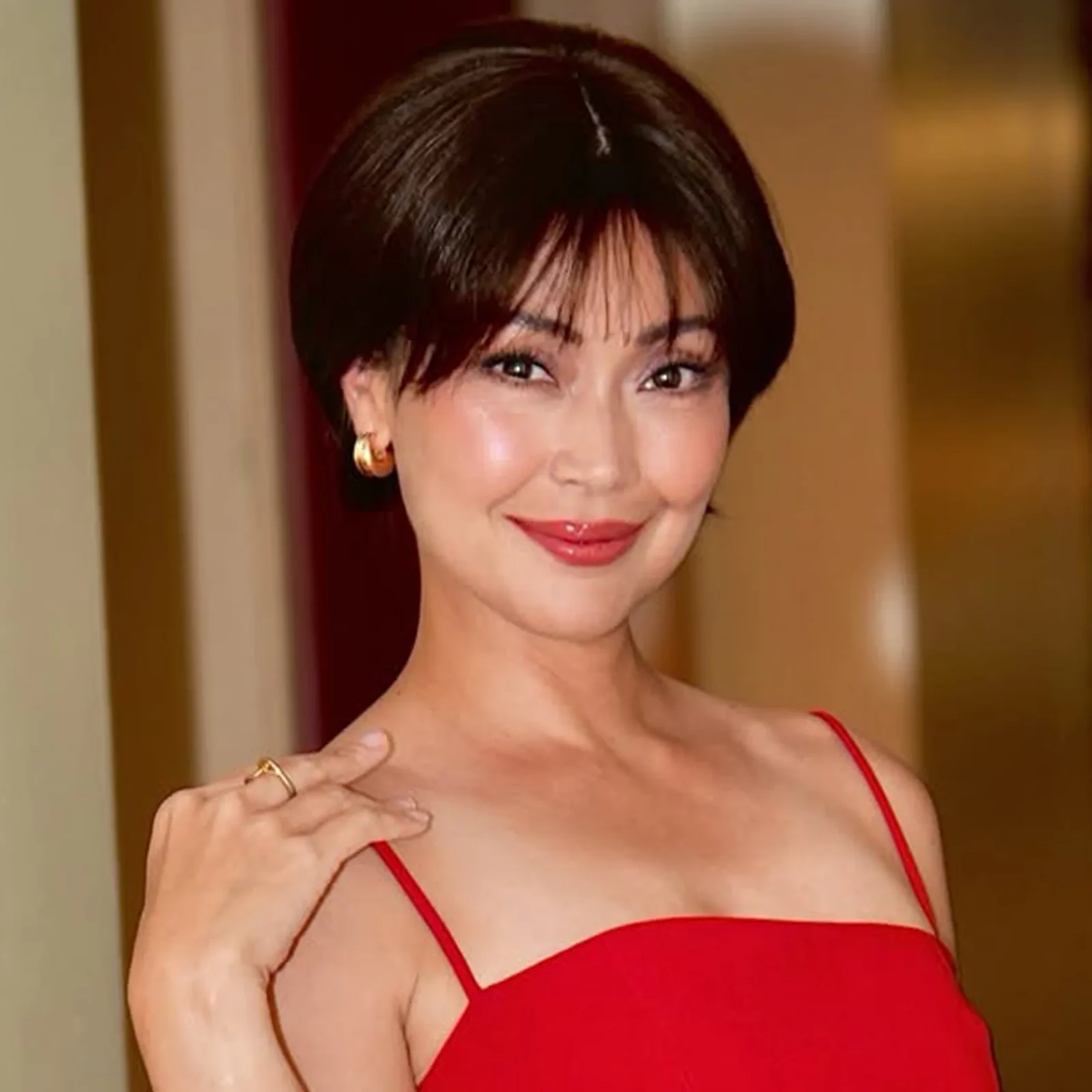 Portrait of a smiling woman with short dark hair wearing a red spaghetti-strap dress indoors, gold earrings visible.