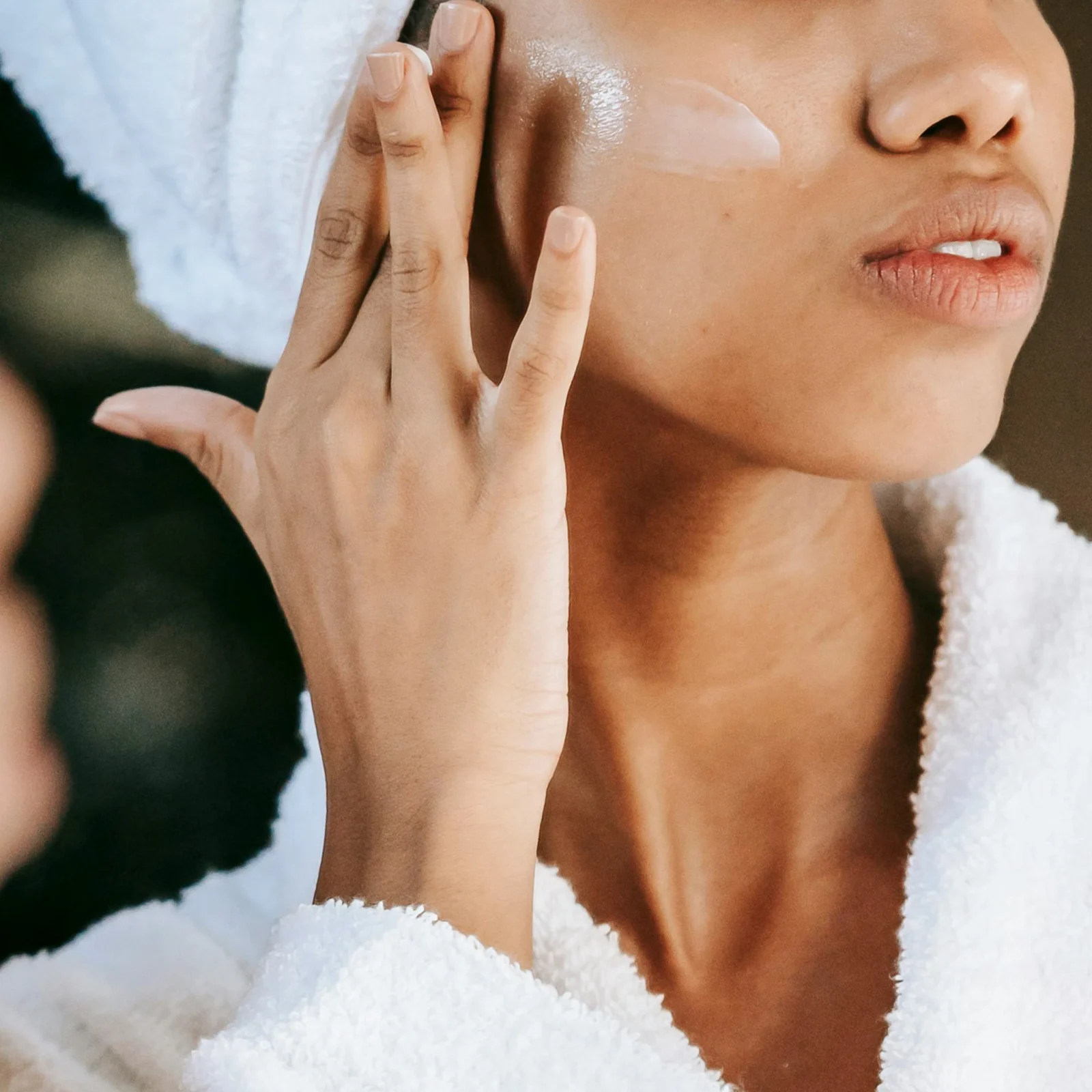 Close-up of a person applying lotion to their cheek while wrapped in a towel.