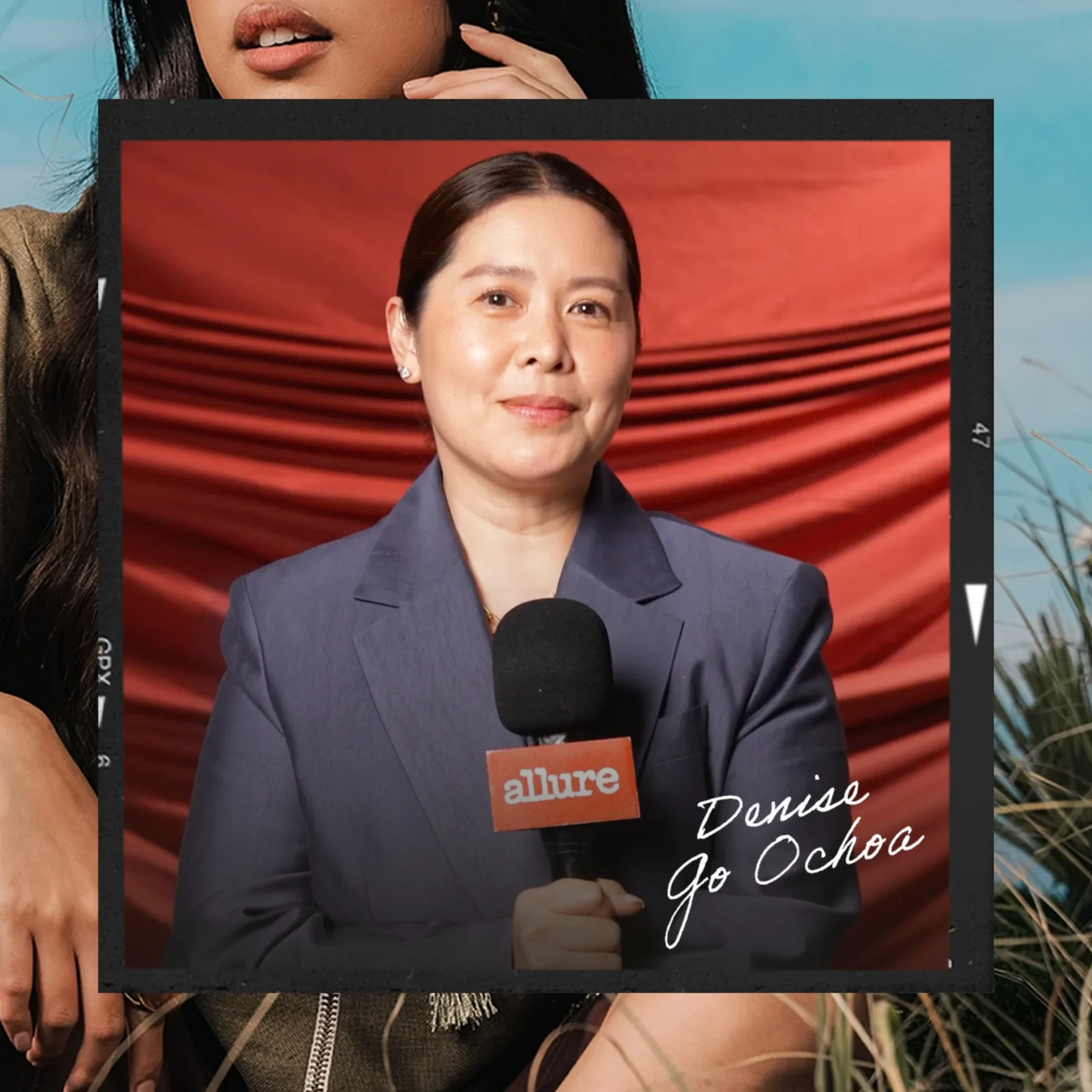 Woman in a blue blazer speaks into a handheld microphone at a media event, with a red wavy backdrop behind her and an Allure badge visible.