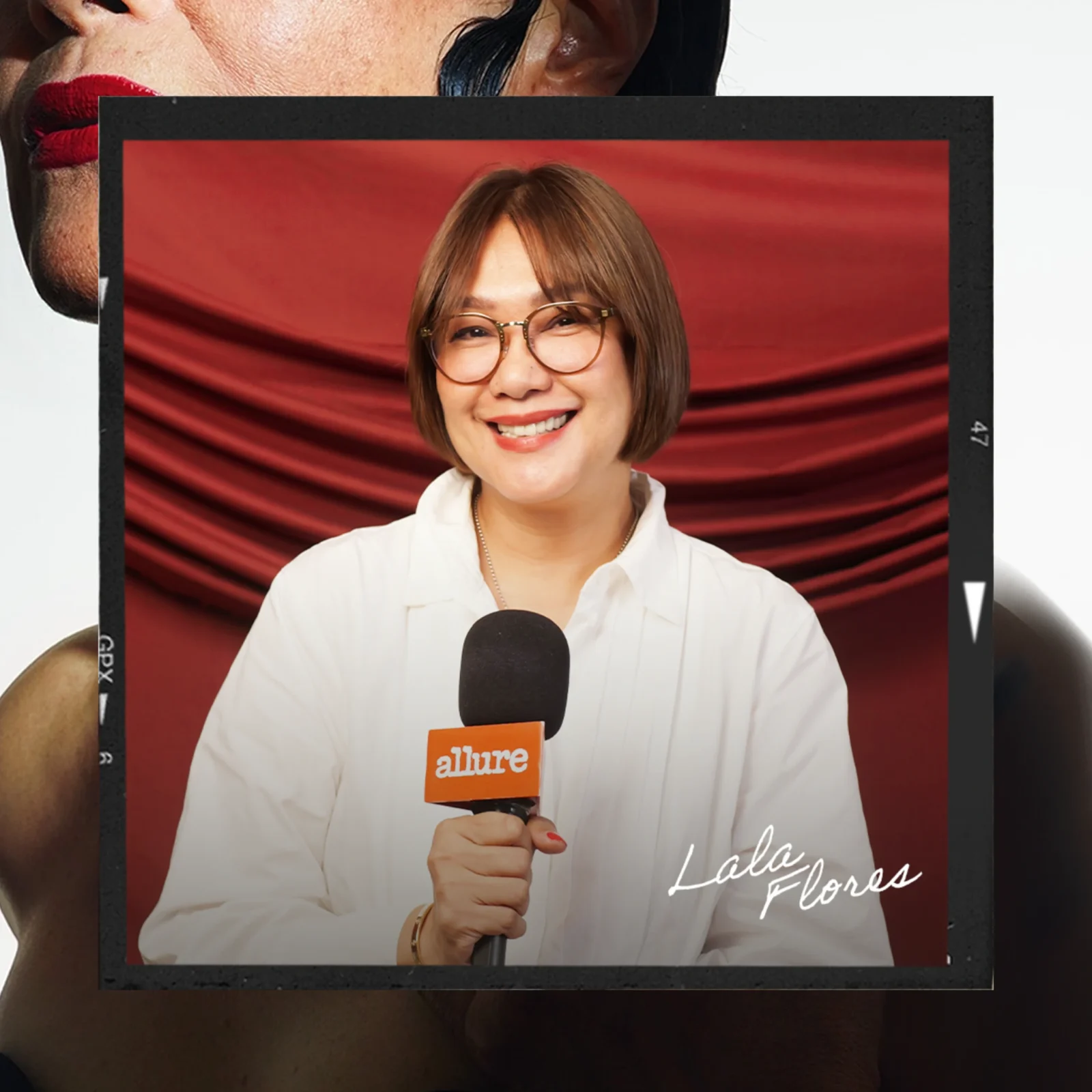 Smiling woman wearing glasses holds a microphone with an Allure badge, in front of a red backdrop during an interview.
