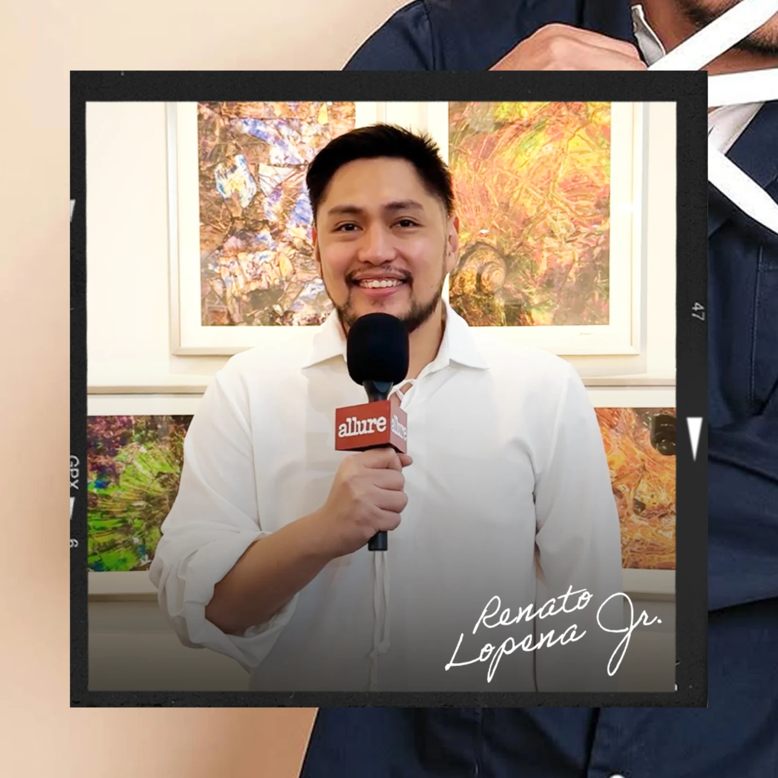 Man in a white shirt speaks into a handheld microphone labeled 'allure' during an interview, with colorful abstract art in the background.