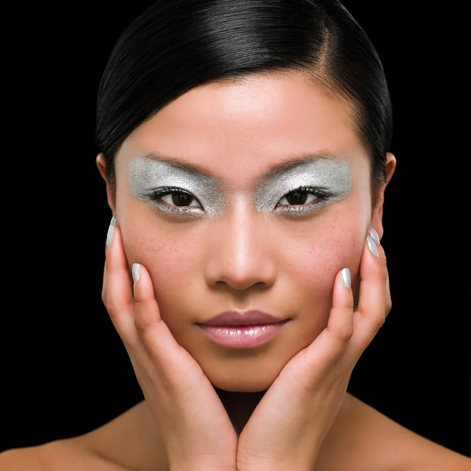 Close-up of a woman with silver eye makeup, hands cupping her face against a black background.